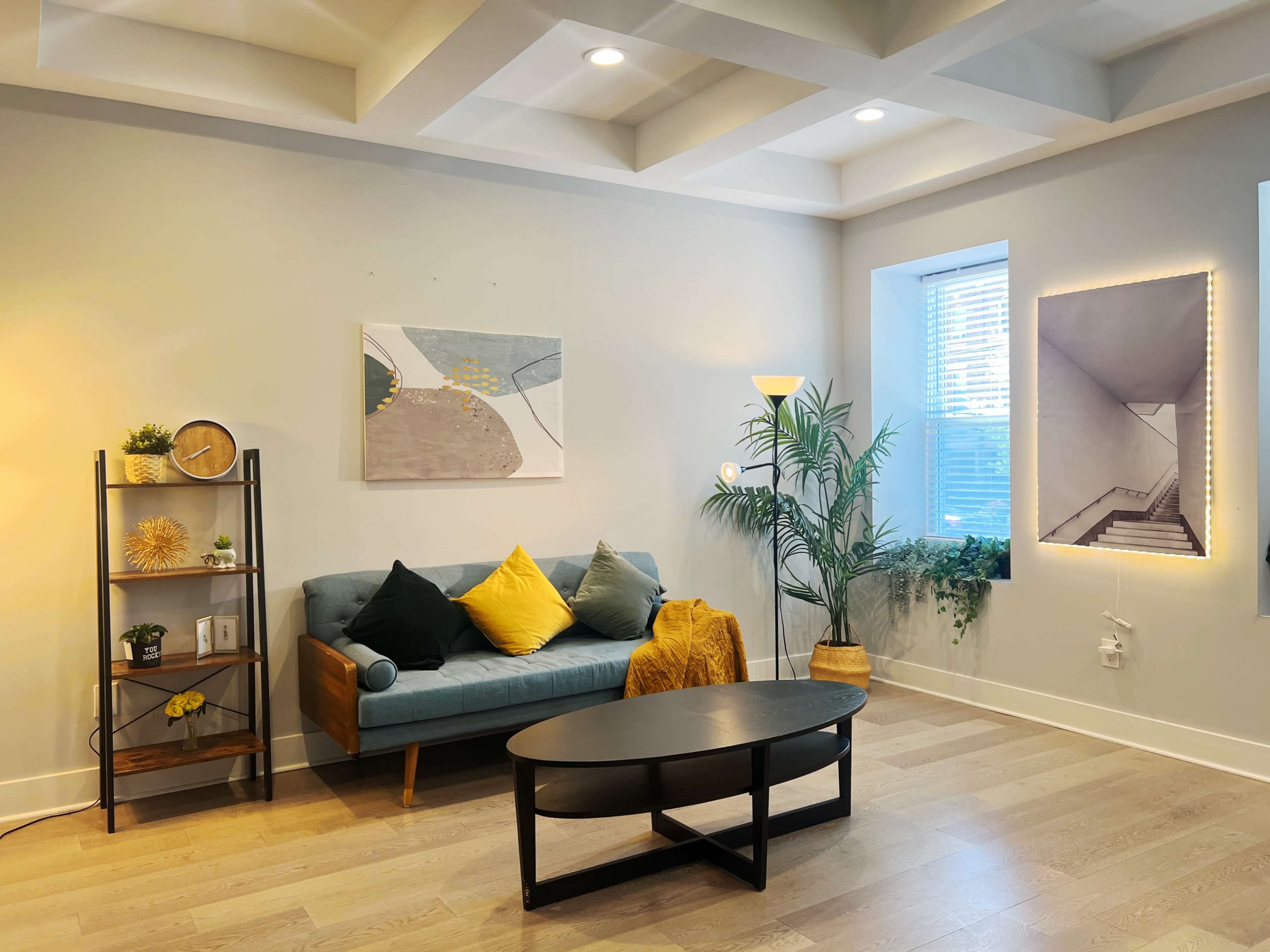 A modern living room features a teal sofa with yellow cushions, a black coffee table, a bookshelf with decorative items, and large windows allowing natural light to illuminate the space.