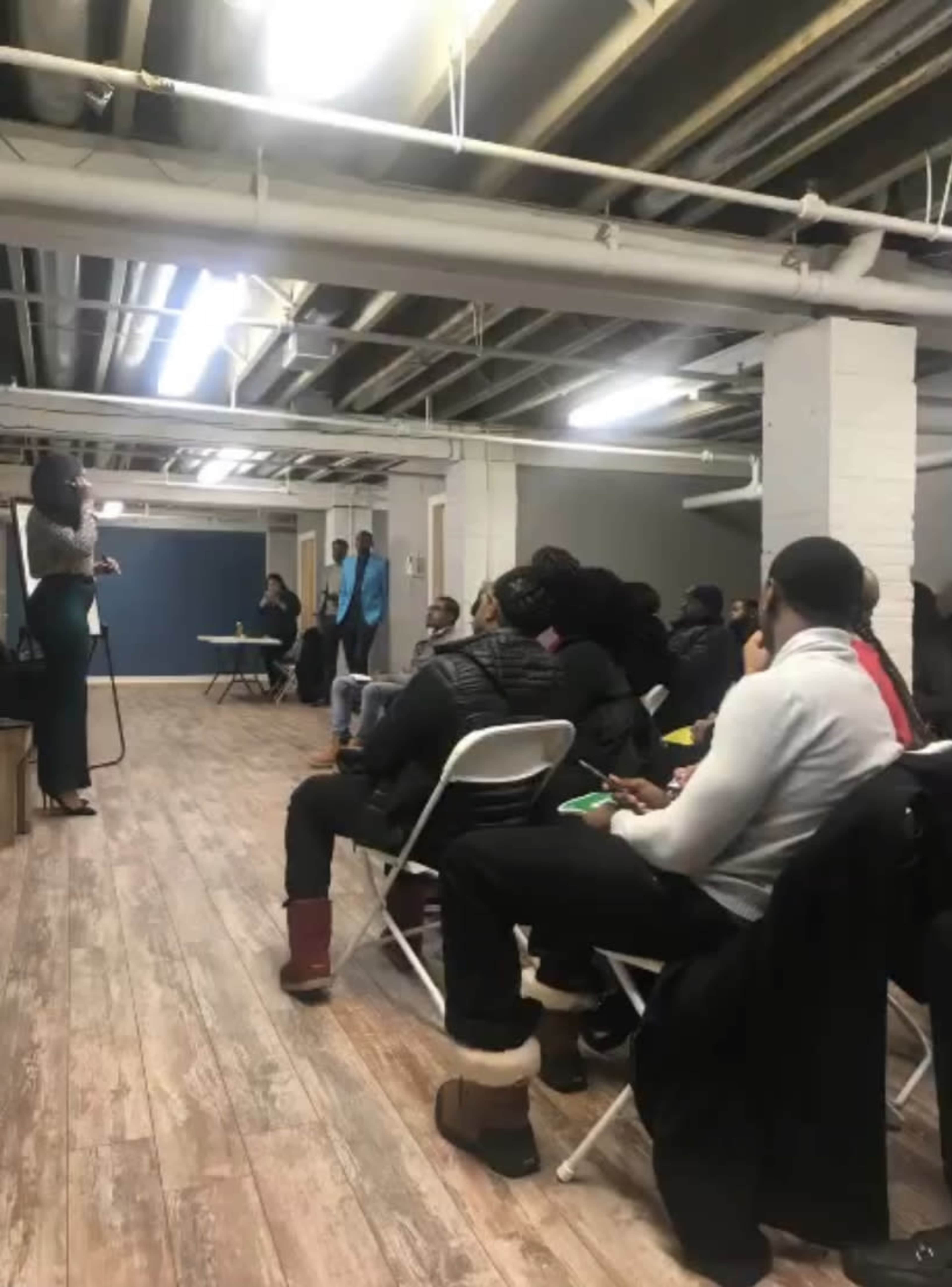 A speaker stands in front of an audience seated in chairs at a gathering in a basement setting.