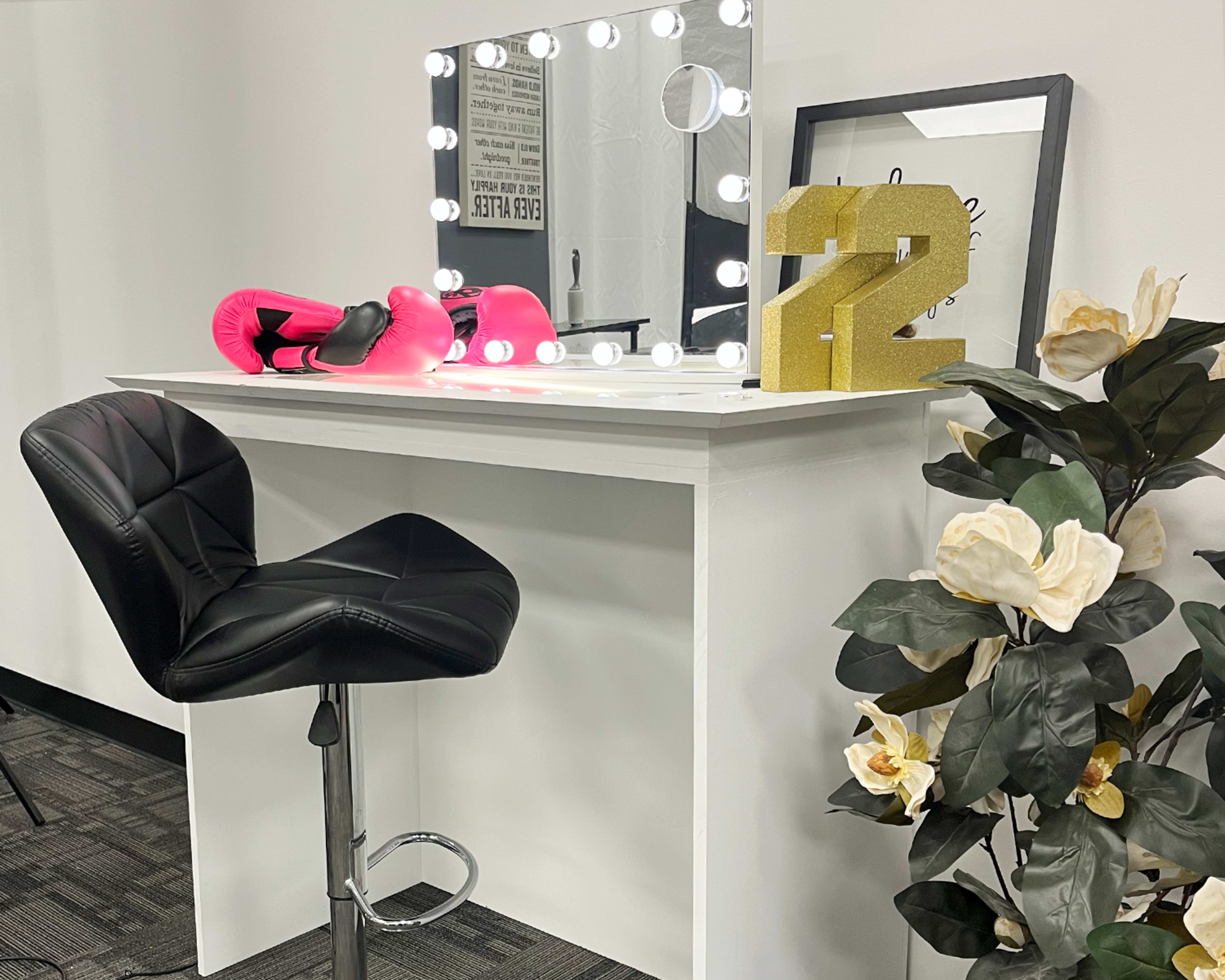 A white vanity table with a large illuminated mirror, a black bar stool, pink boxing gloves, a golden number 12 decoration, and a potted plant.
