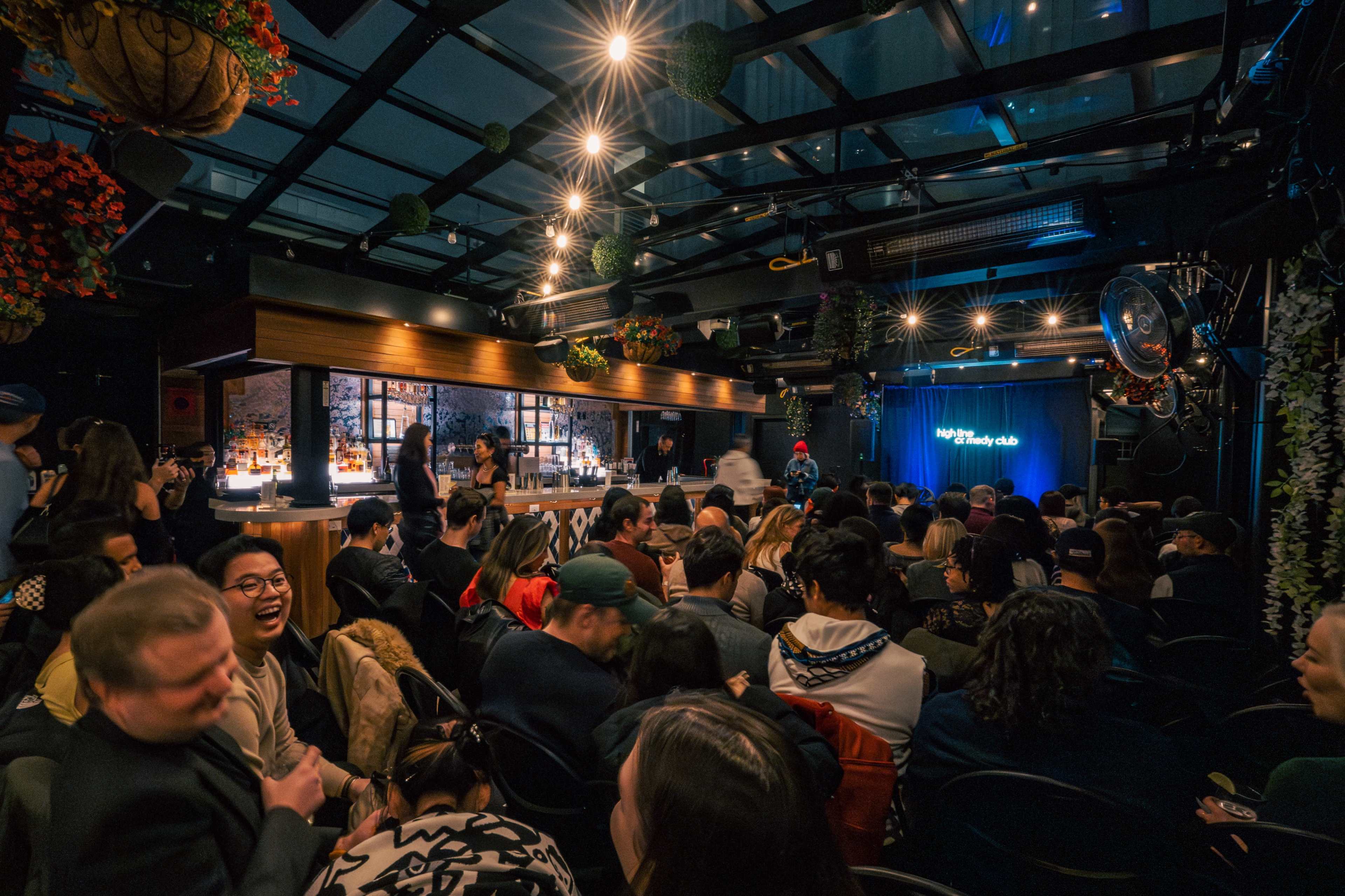 A crowded comedy club features an audience seated in anticipation, with a stage set for a performance in the background.