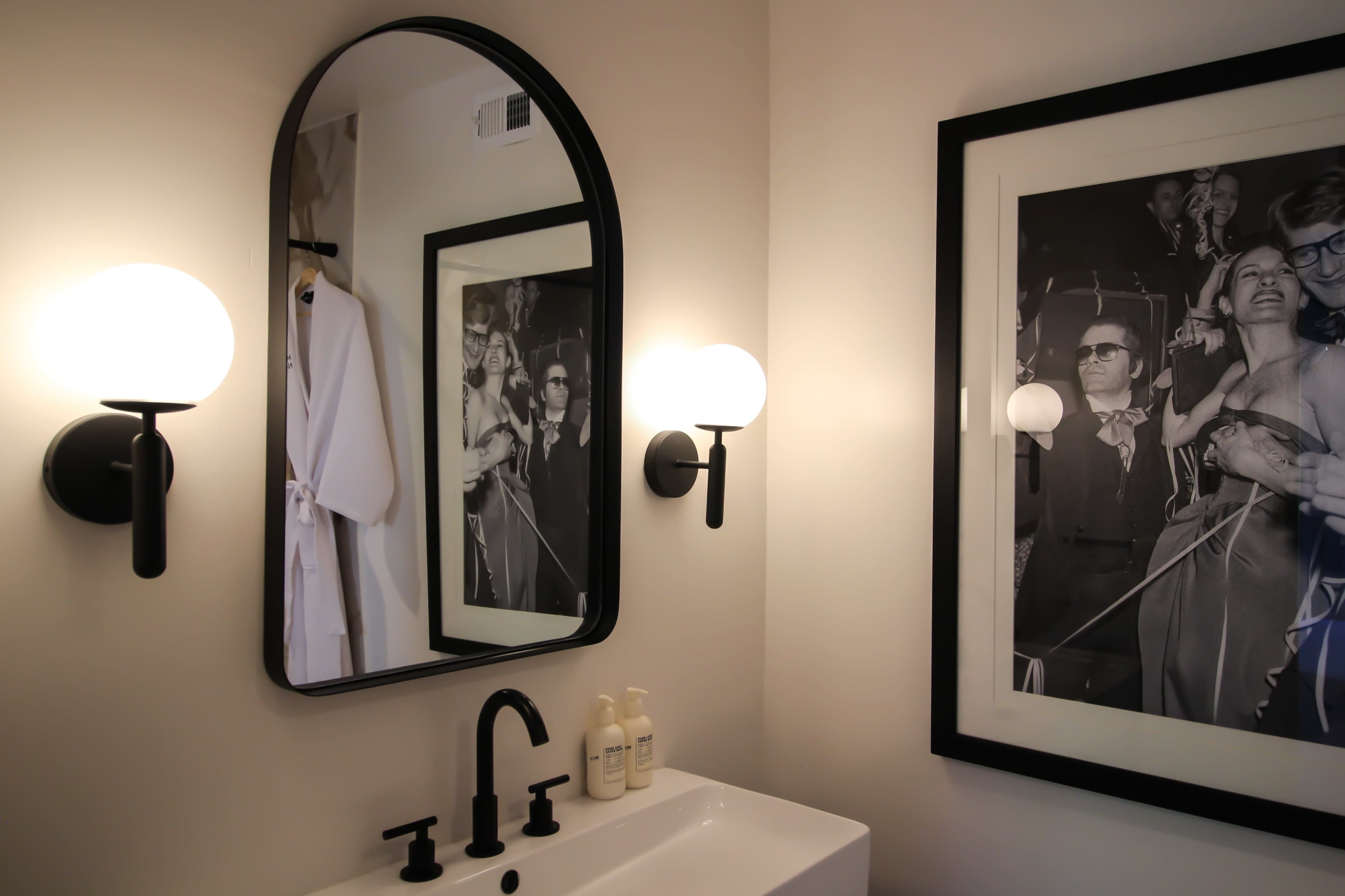 A modern bathroom with a sleek sink, wall-mounted faucets, a round mirror, and a large framed black-and-white photograph of people at an event.