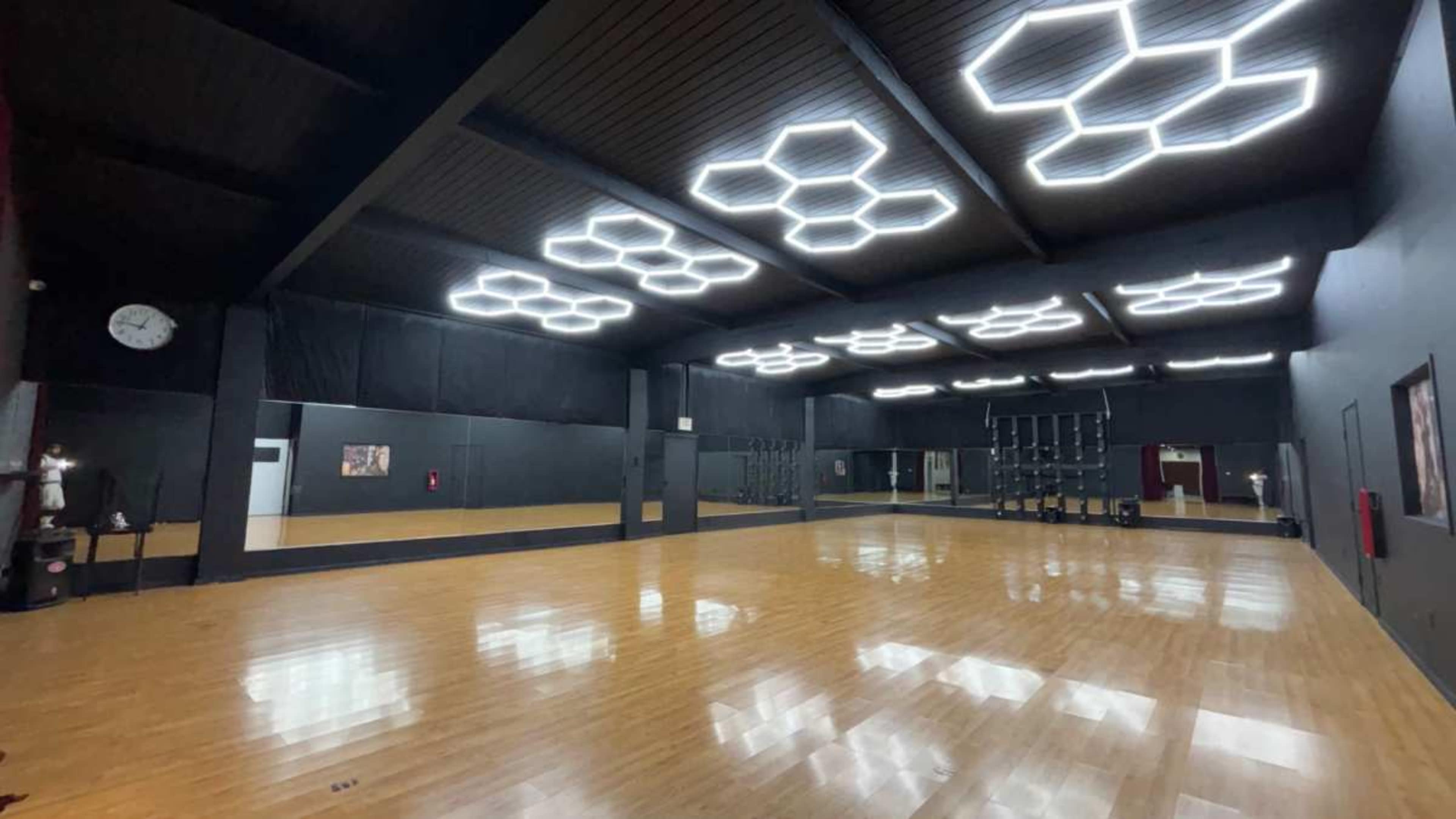 The image shows a spacious dance studio with a polished wooden floor, mirrored walls, and modern hexagonal ceiling lights.