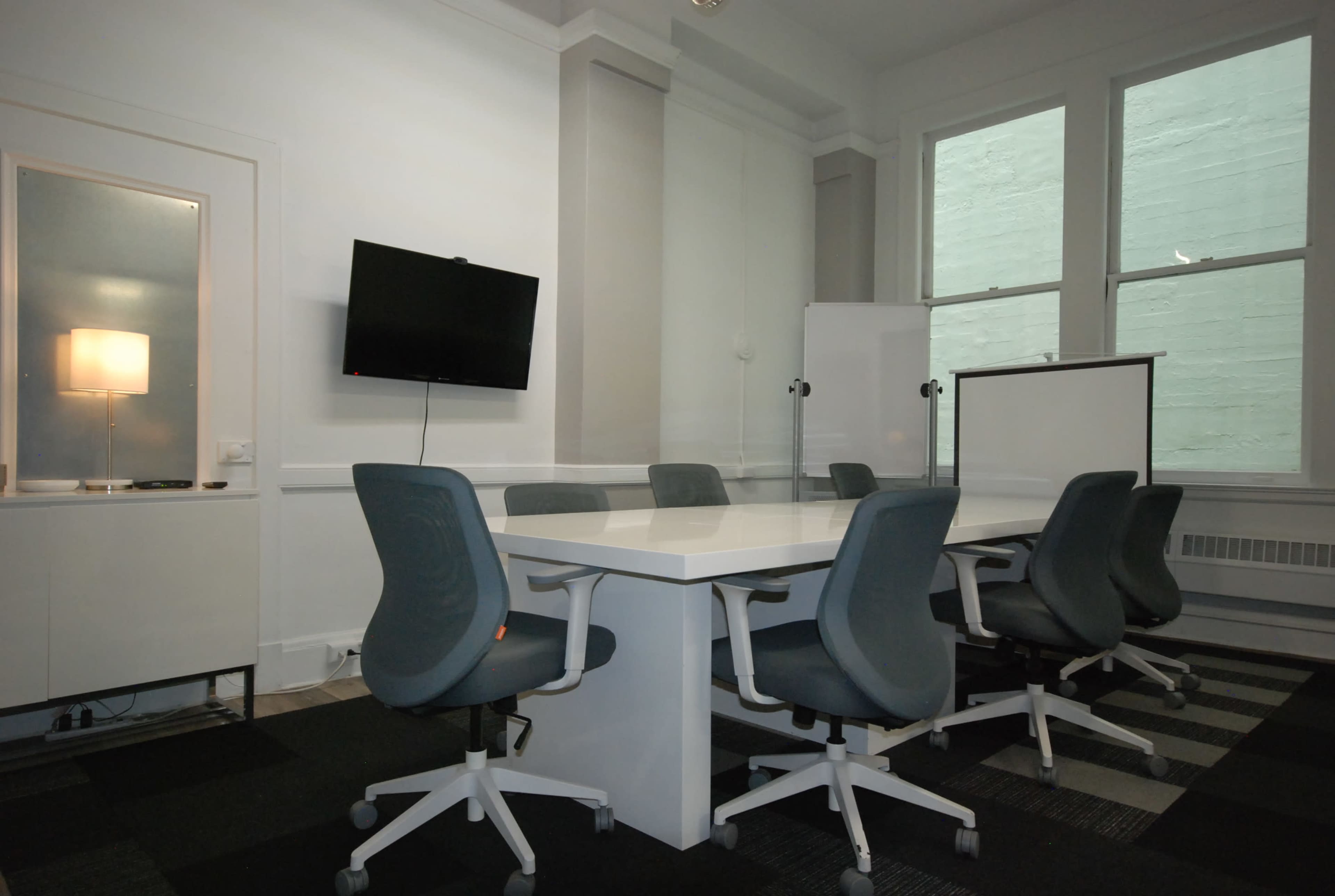 A modern conference room features a white table surrounded by gray chairs, with a television mounted on the wall and a large window providing natural light.