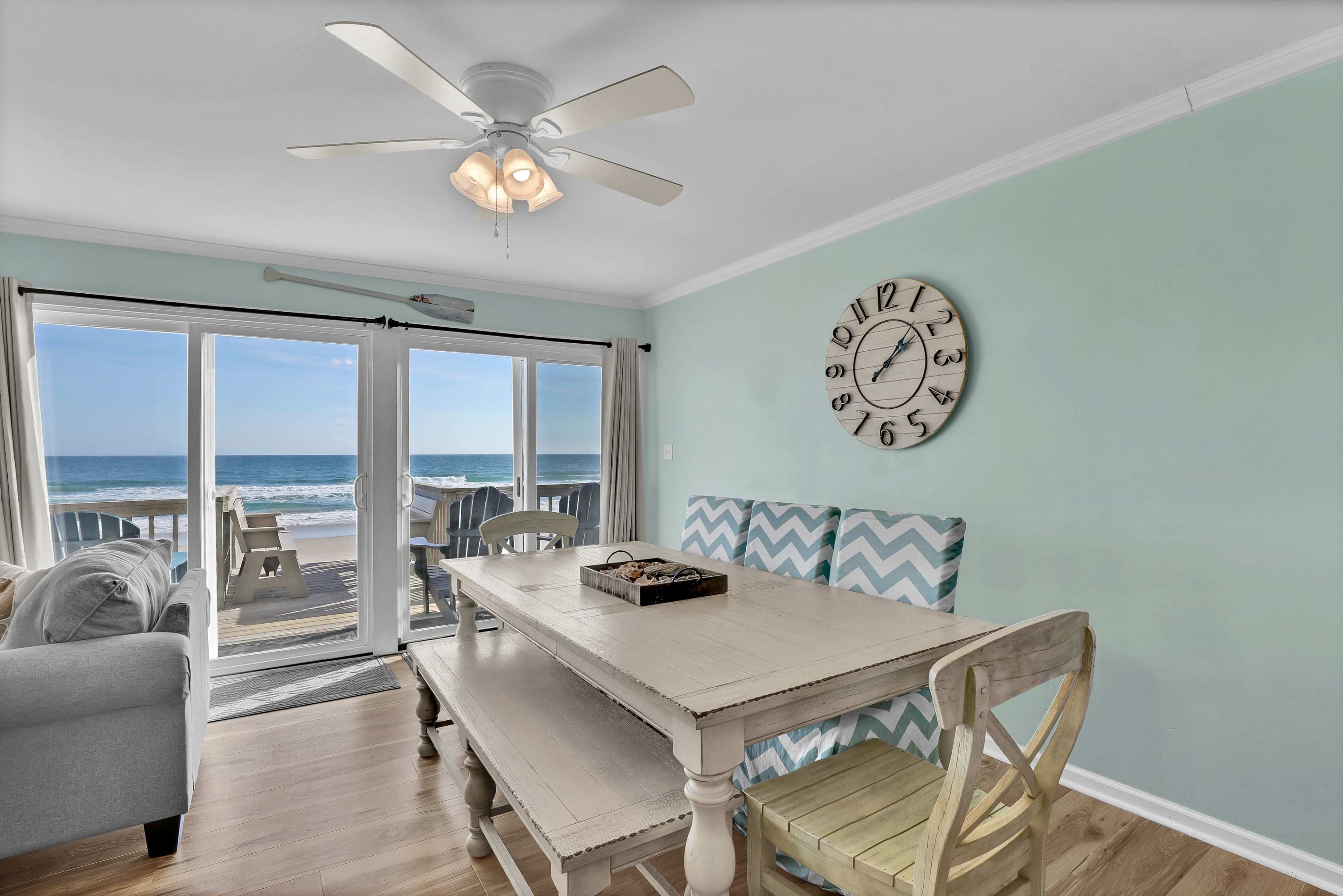 The image shows a dining area with a light-colored table and chairs facing large sliding glass doors that open to a view of the ocean and beach.