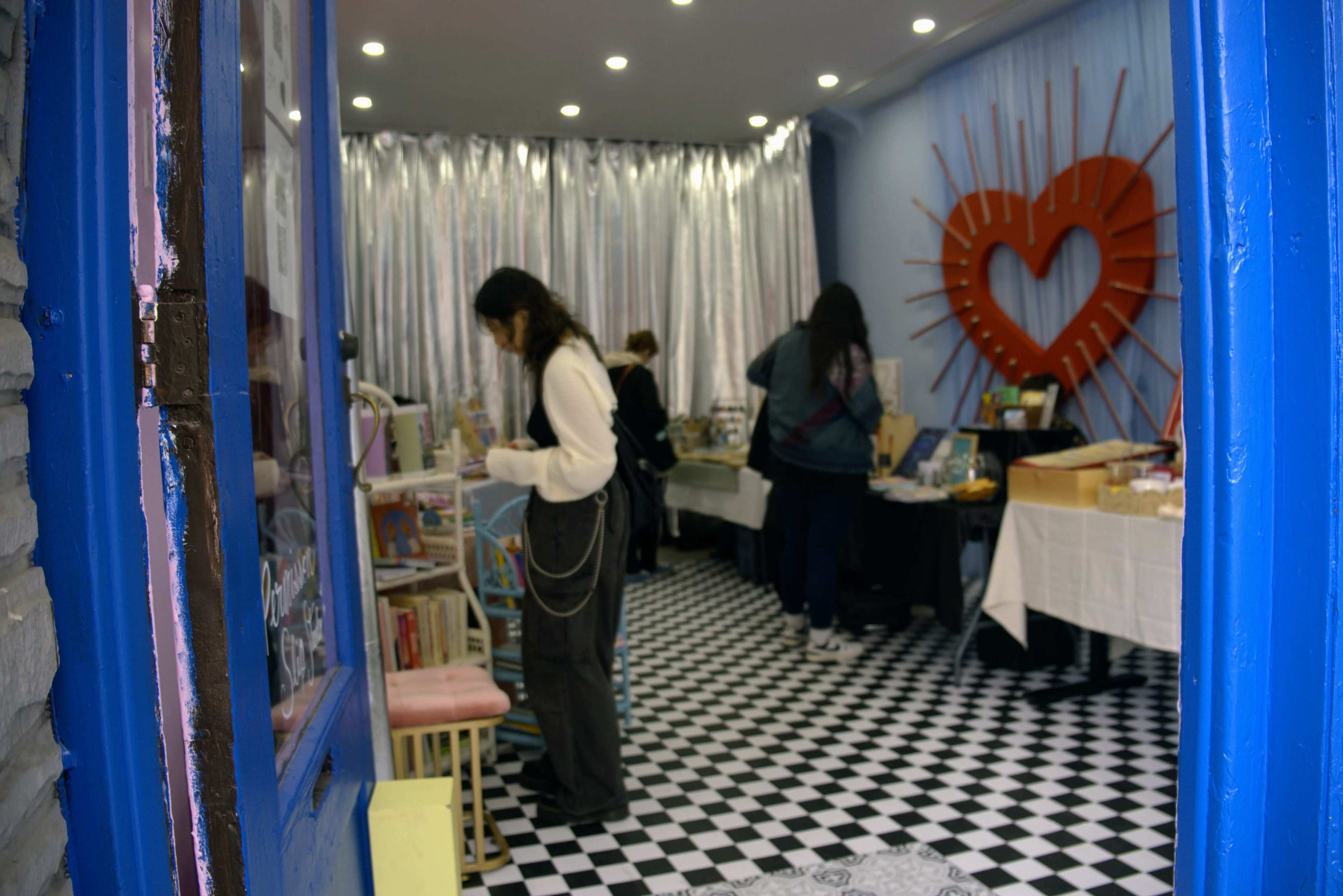 The image shows a boutique with black and white checkered flooring, where several people browse through items on display tables amidst a backdrop of silver curtains and a large red heart decoration.