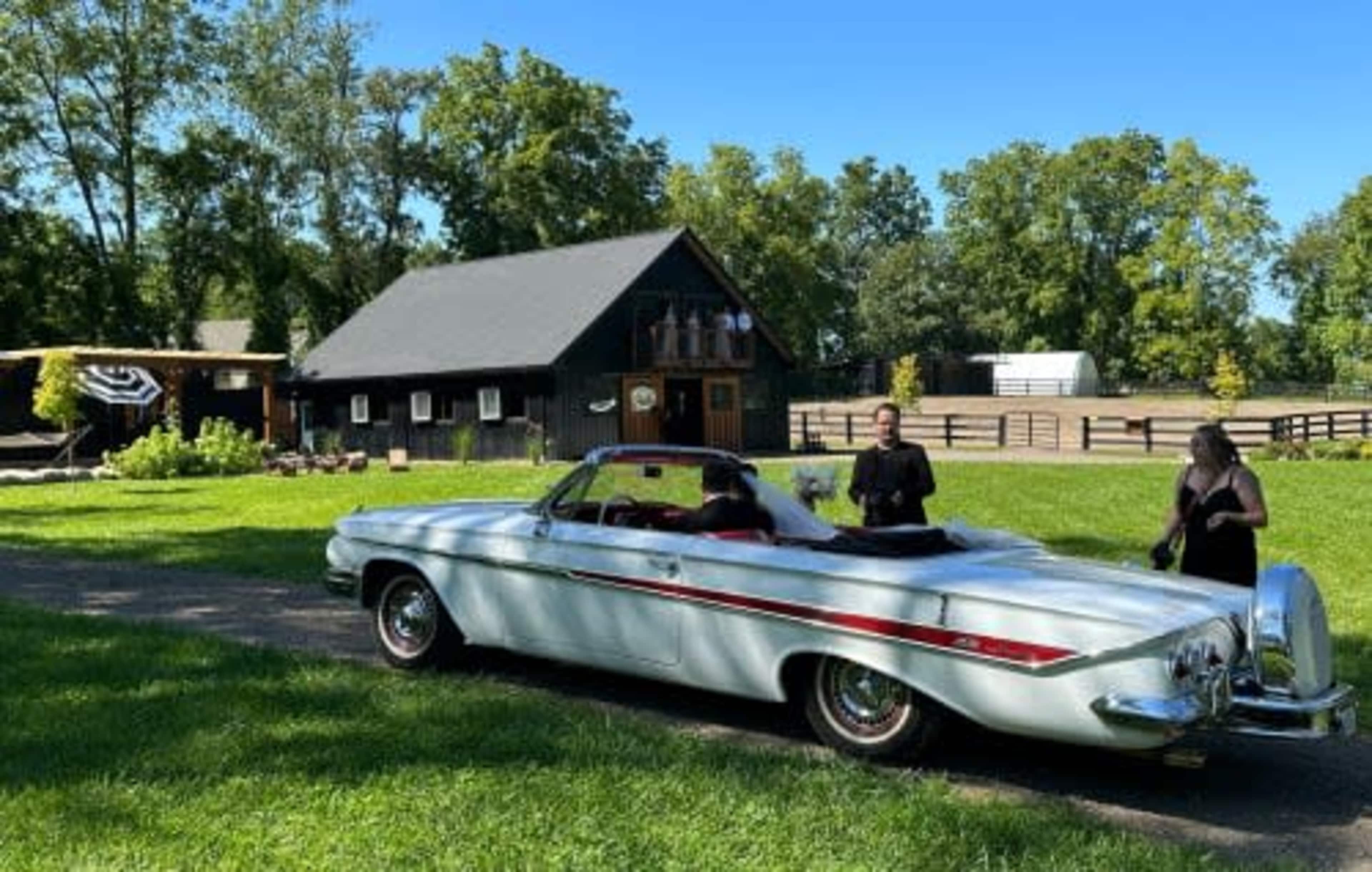A classic convertible car is parked on a gravel path in front of a black barn while two individuals stand beside it on a lush green lawn.