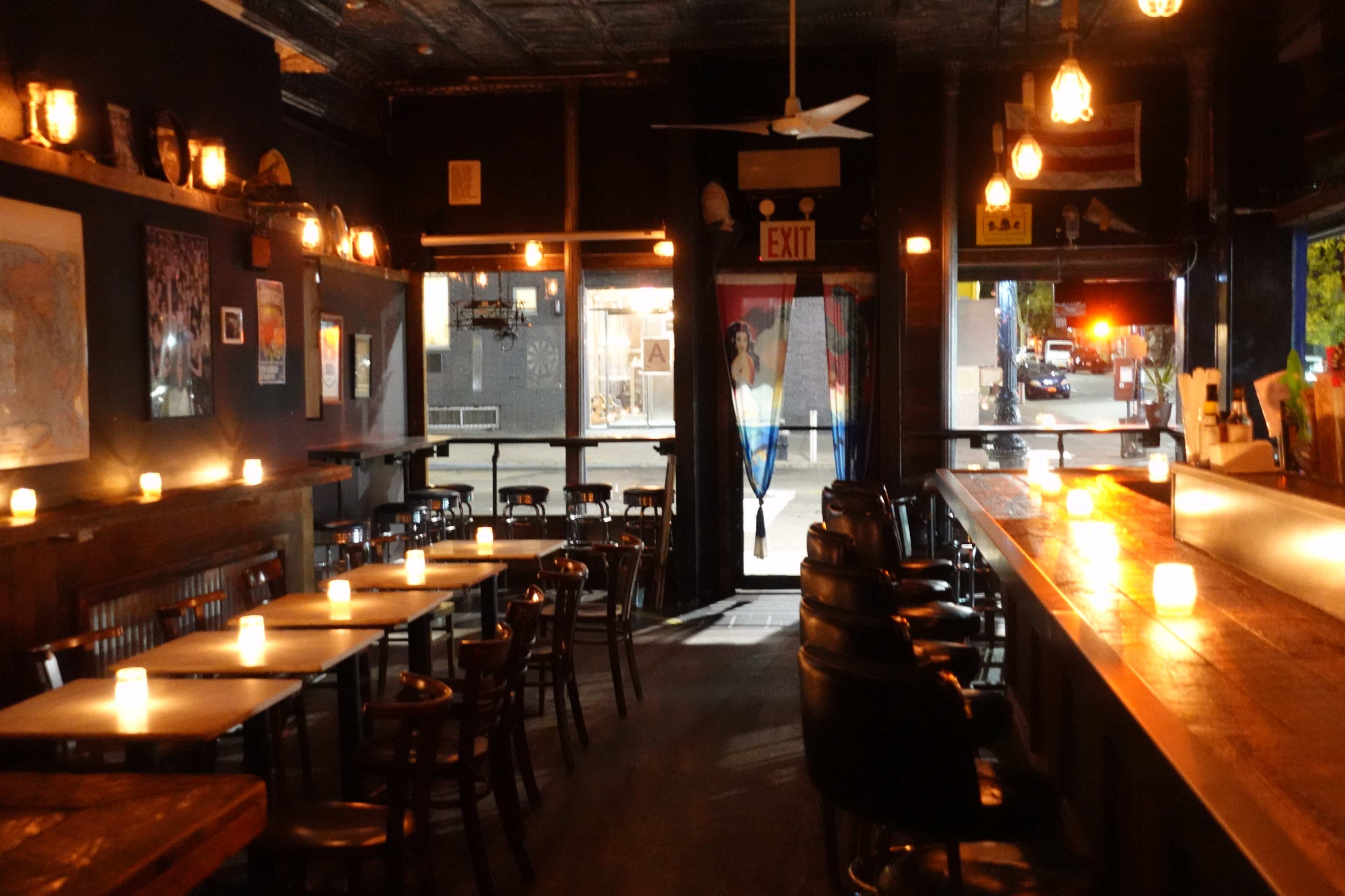 The image shows the interior of a dimly lit bar with wooden tables and chairs, candles on the tables, and an open door leading to a street outside.