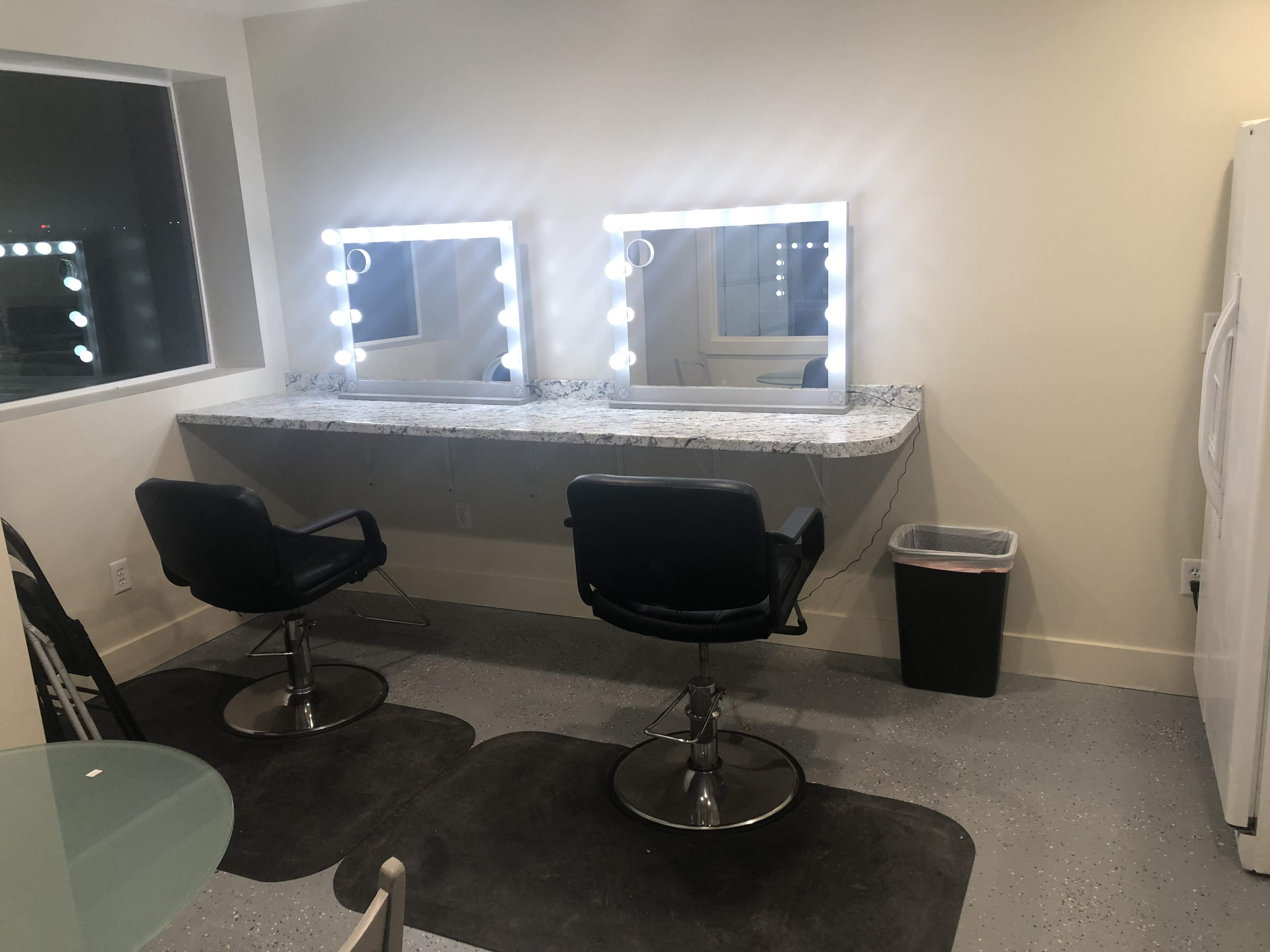 A modern salon space featuring two black chairs in front of illuminated mirrors, with a counter and a trash can nearby.