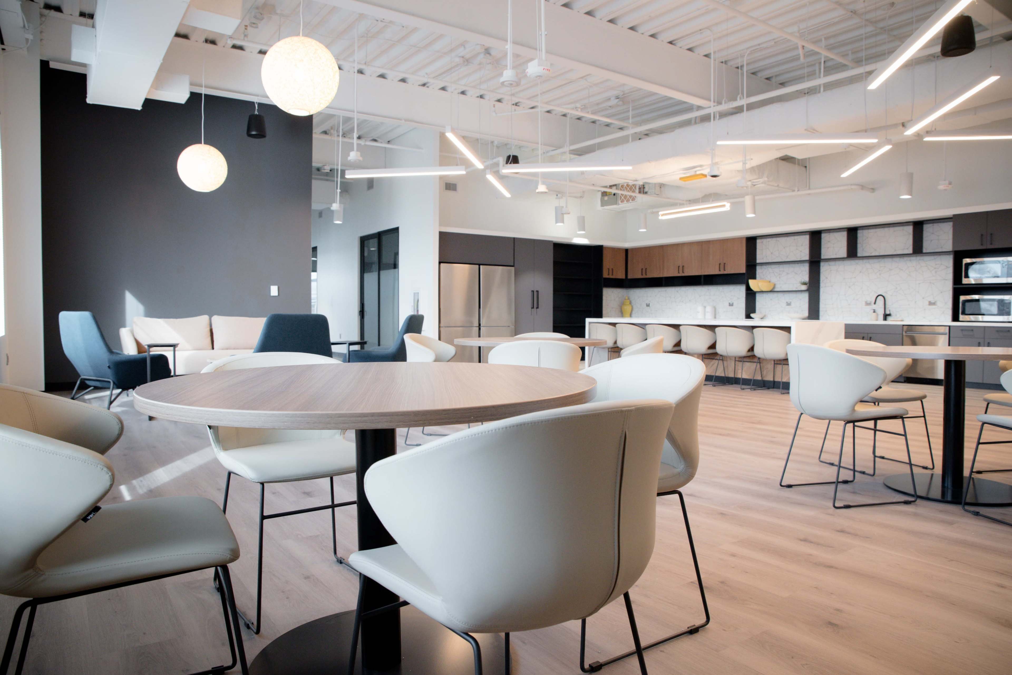 The image shows a modern office break room with a dining table, stylish chairs, and a kitchenette in the background.