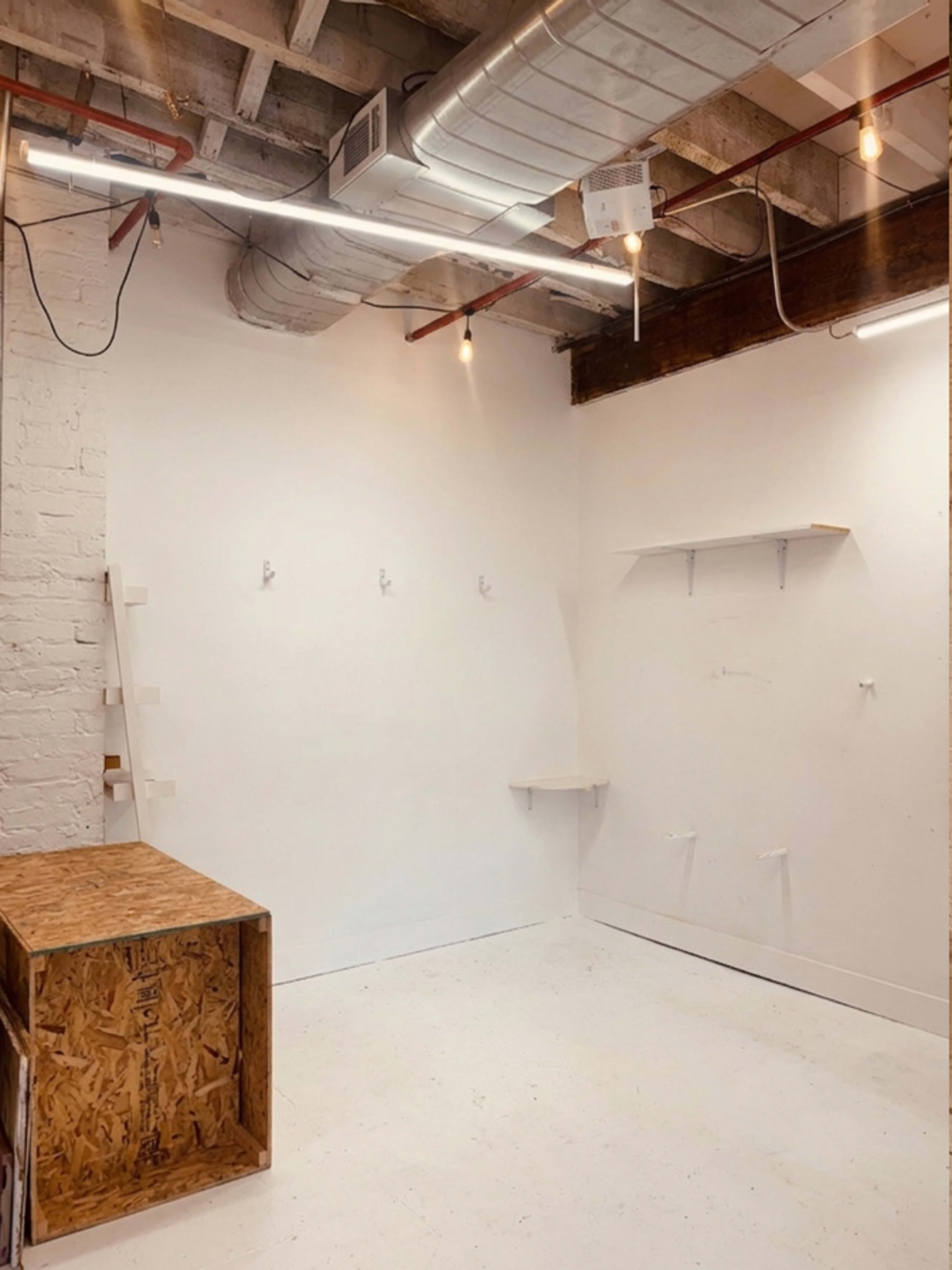 The image shows a minimalistic room with white walls, a wooden countertop or box, several hooks mounted on the wall, and exposed ceiling infrastructure.