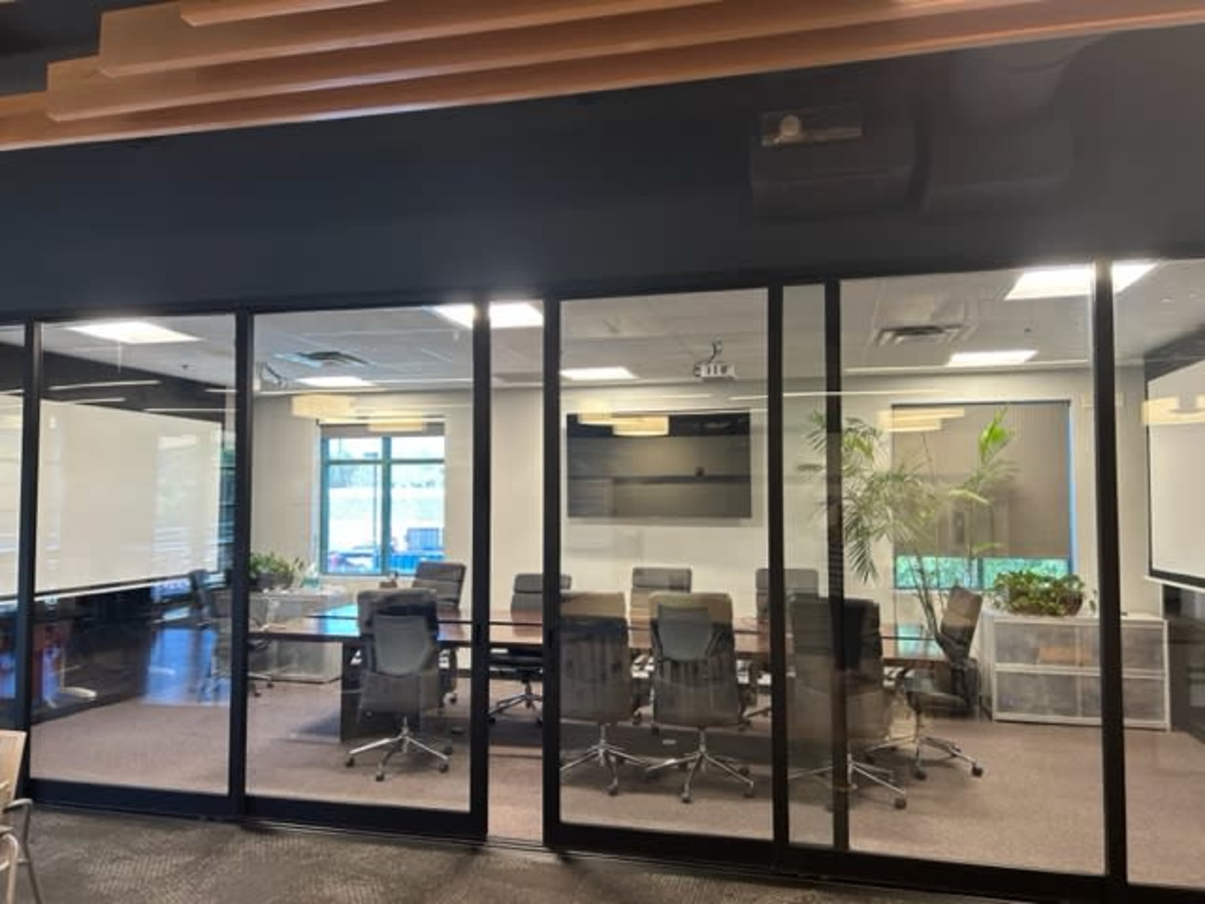 A glass-walled conference room with a long table, office chairs, and a large screen mounted on the wall.