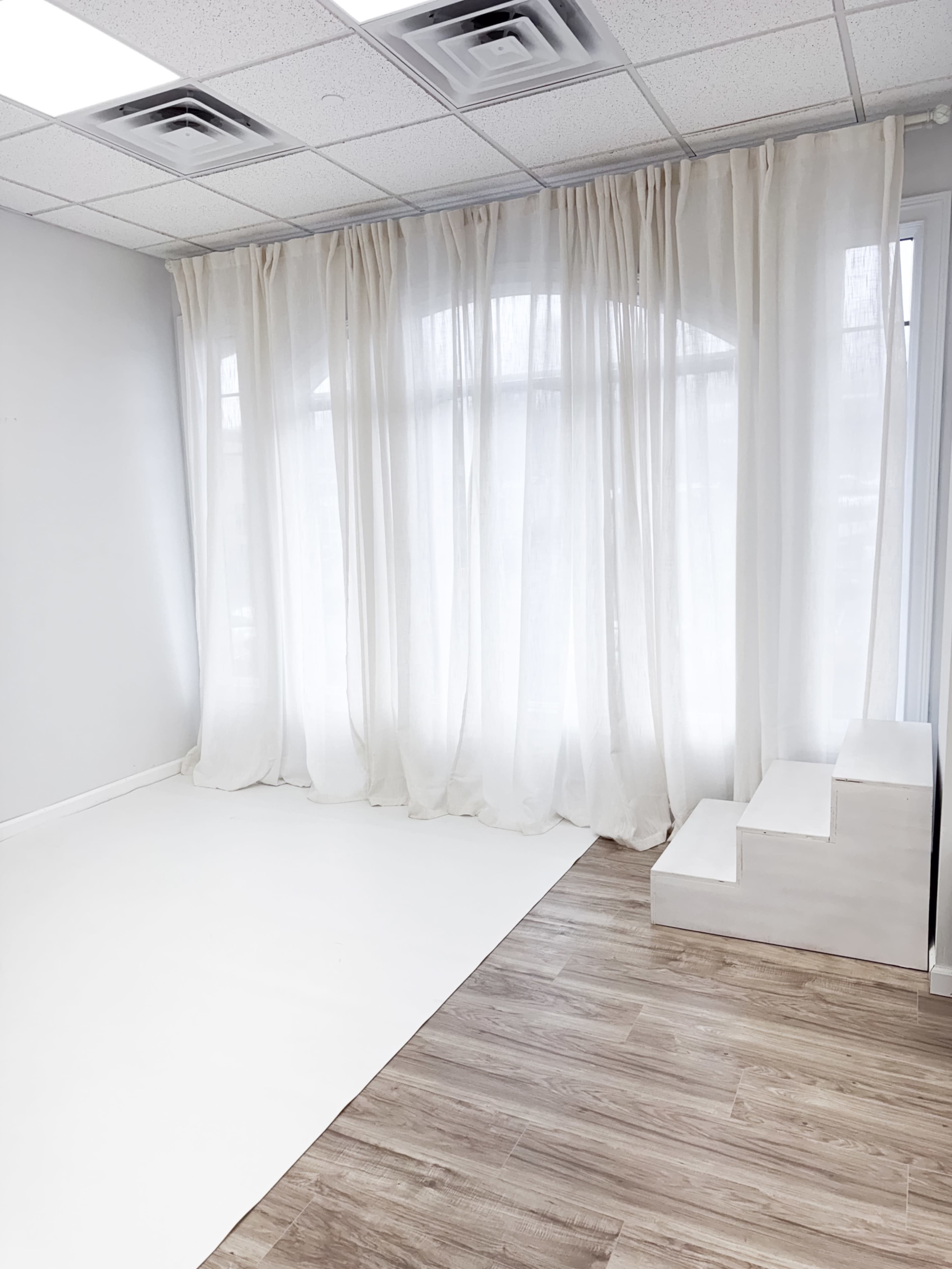 The image shows a minimalist room with light-colored walls, a white floor, a set of three stairs, and sheer white curtains covering large windows.