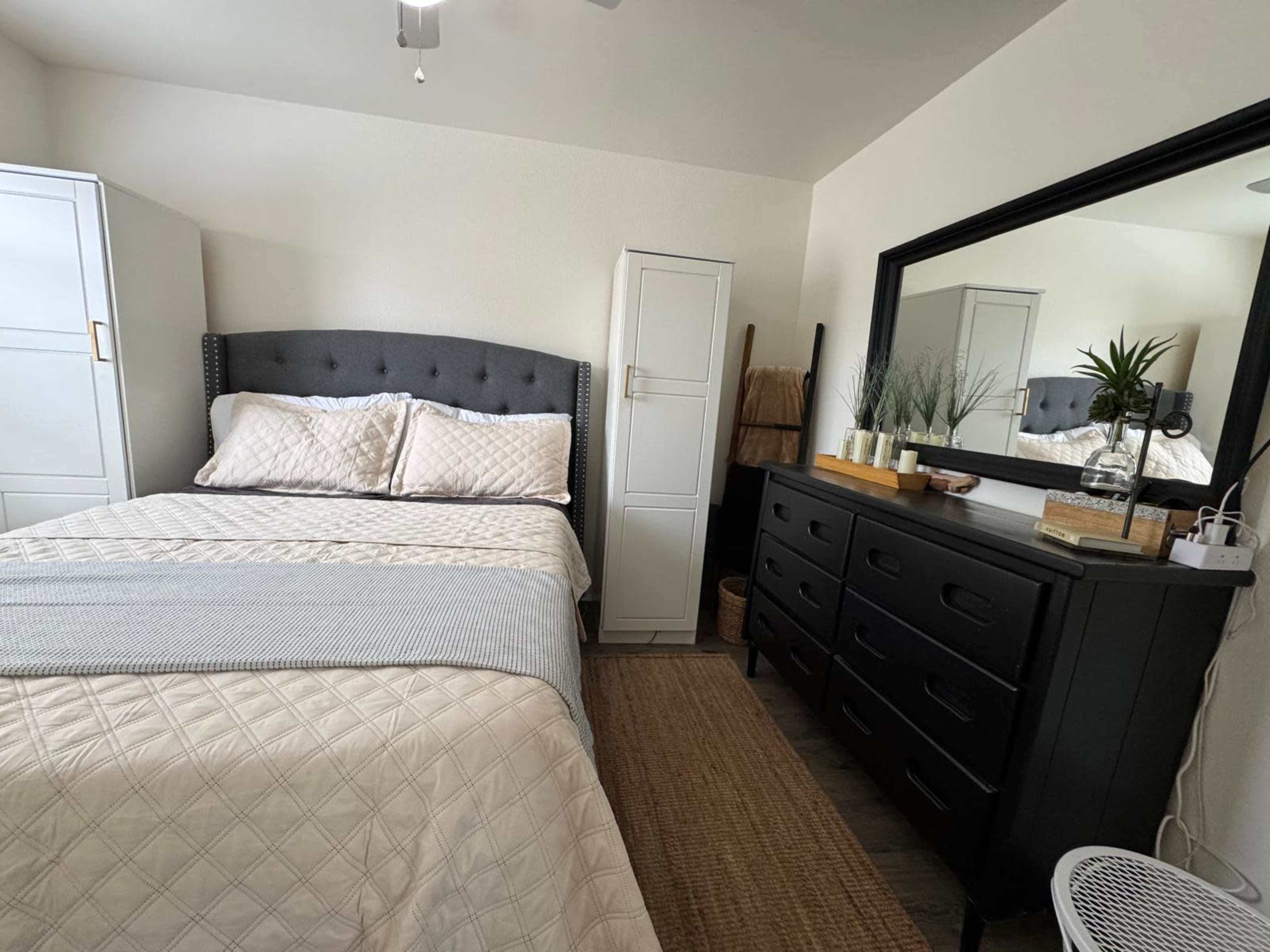 The image shows a neatly arranged bedroom featuring a bed with a gray headboard, a dresser, a mirror, and a wardrobe.