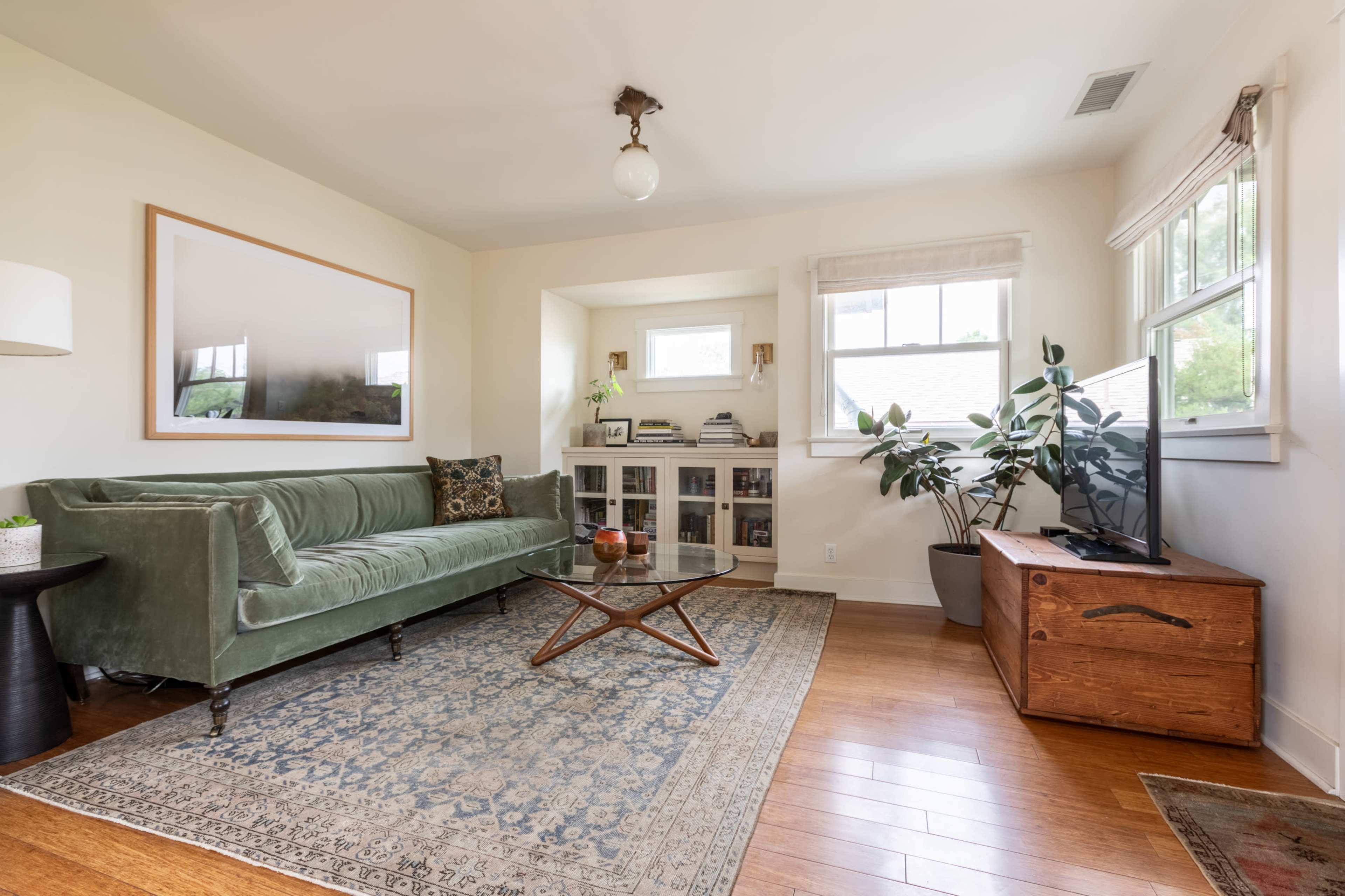 The image shows a light-filled living room featuring a green velvet sofa, a wooden coffee table, and a corner bookshelf.