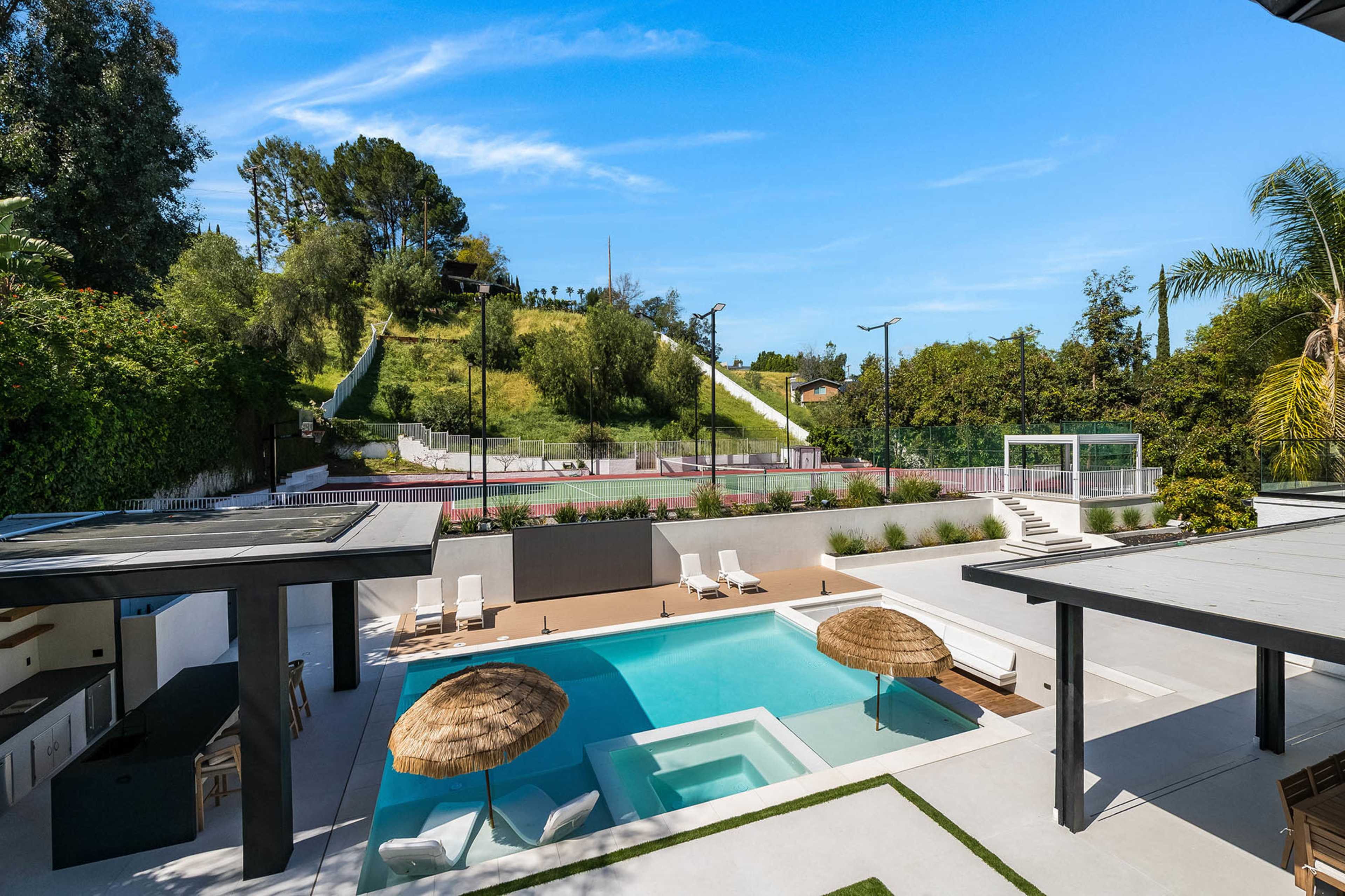 The image shows a modern backyard featuring a swimming pool with two thatched umbrellas, surrounded by a landscaped area and tennis courts in the background.