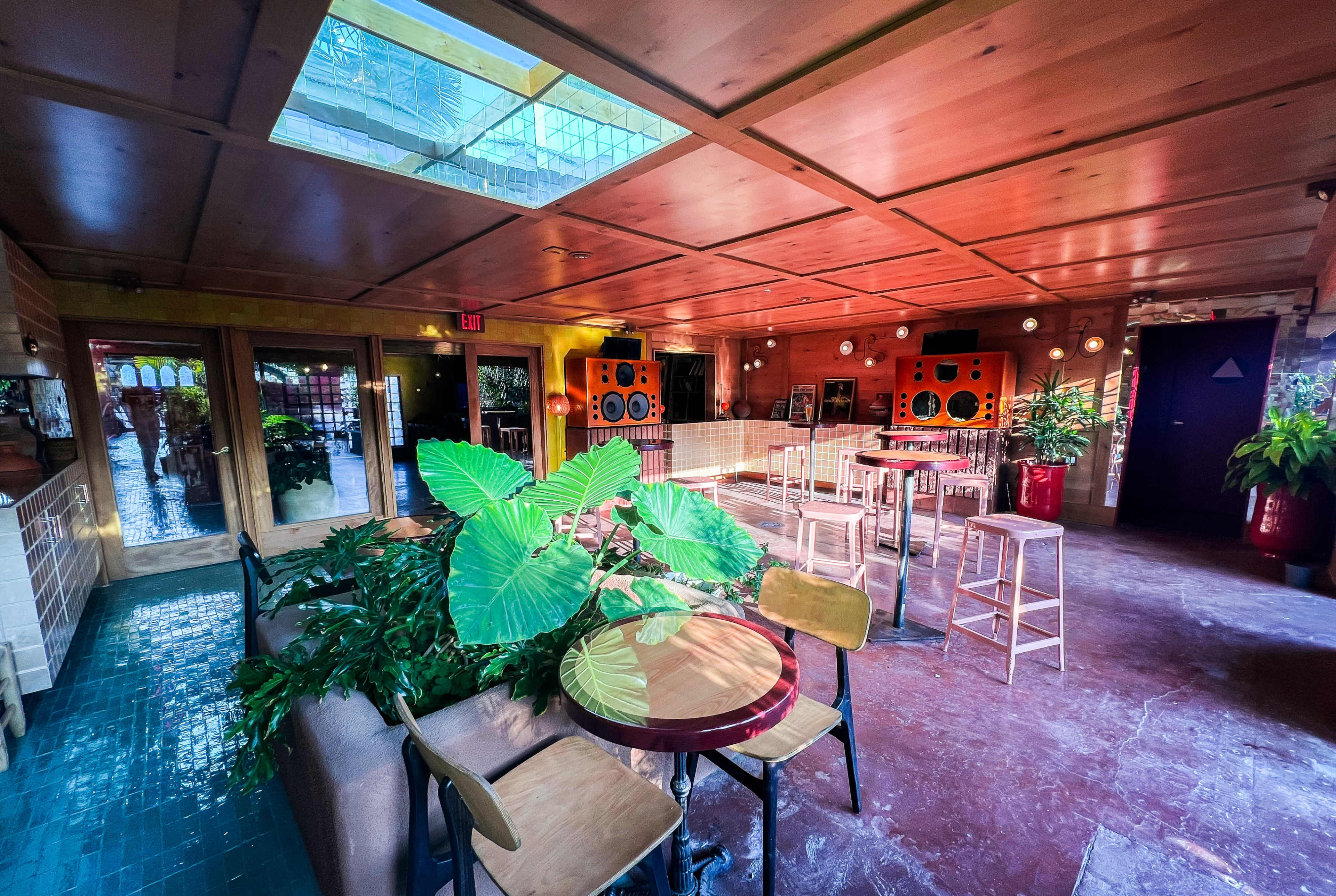 The interior of a colorful, spacious venue features wooden beams, green plants, and a mix of seating arrangements.