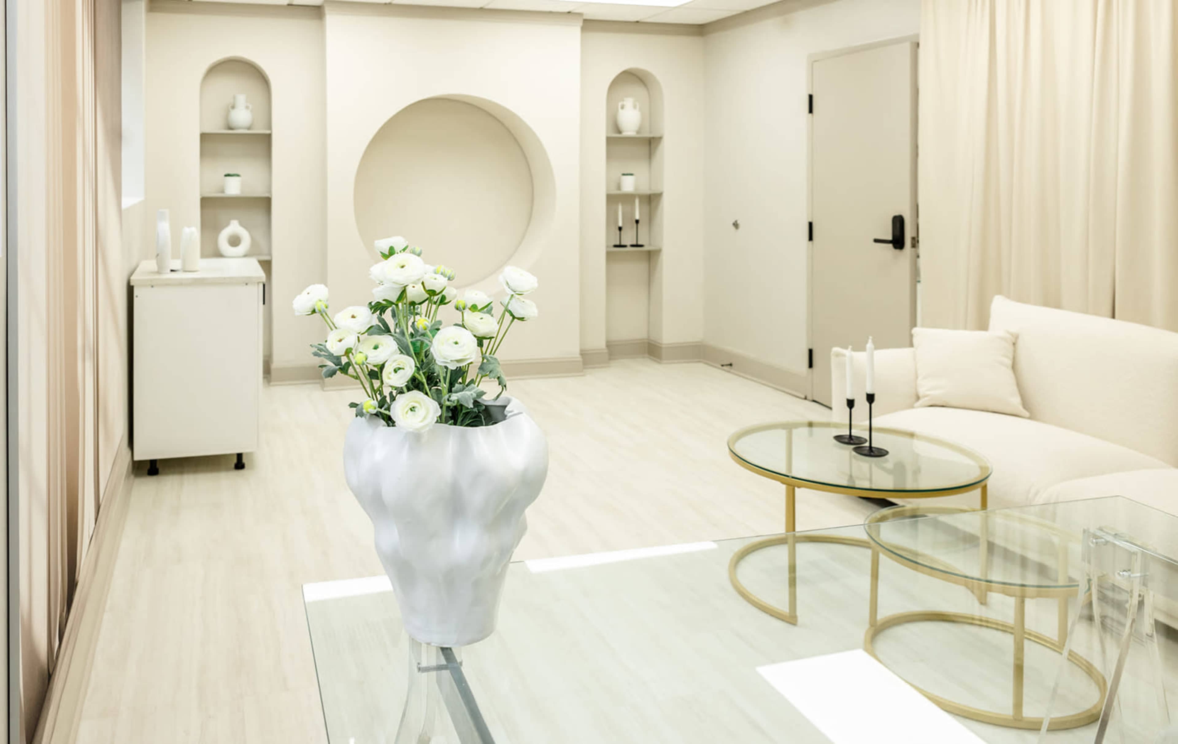 A modern, minimalistic room features a white sofa, glass tables, and a vase of flowers, with light-colored walls and a circular niche in the background.