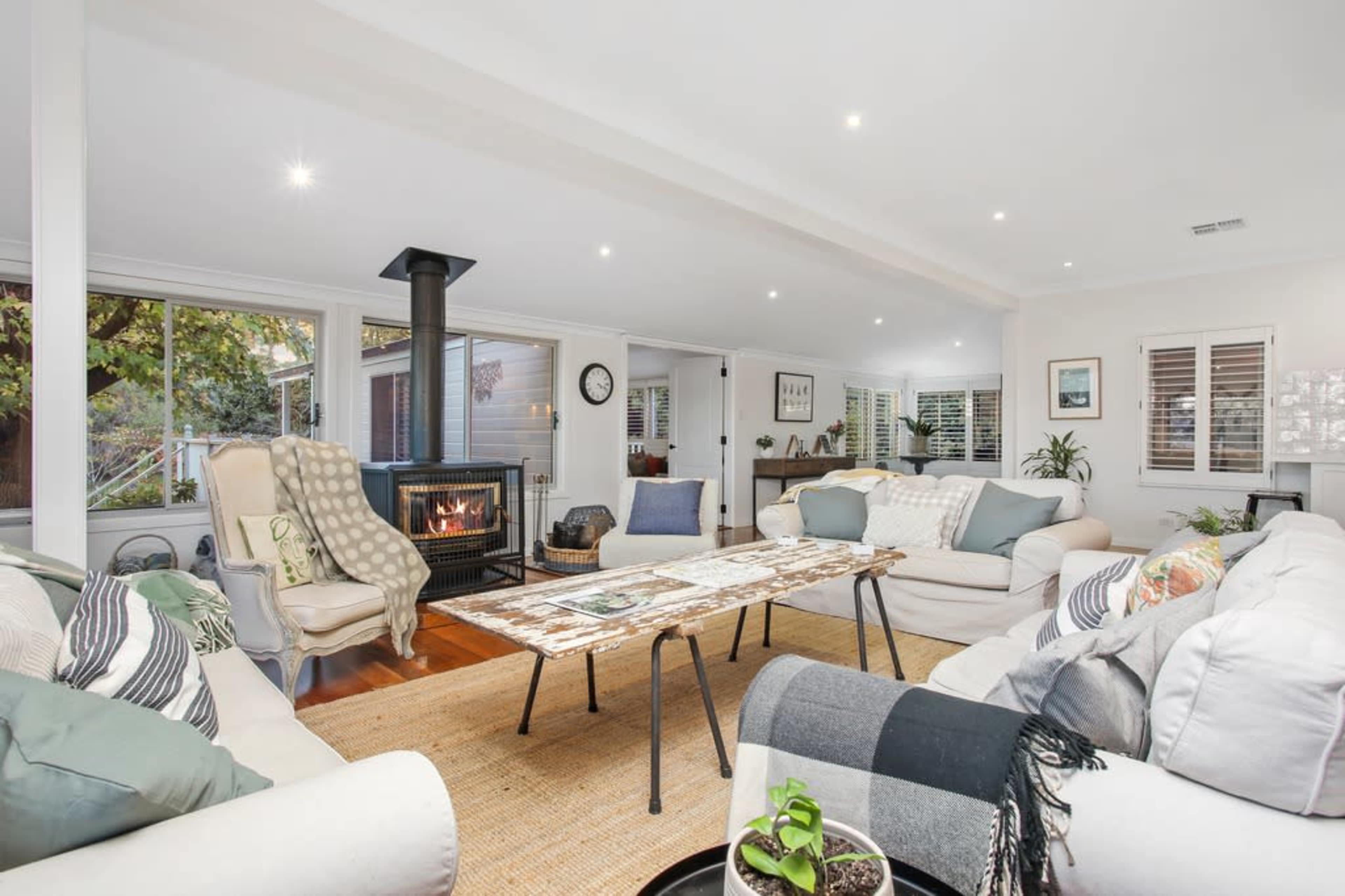 The spacious living room features a central coffee table surrounded by multiple sofas and a fireplace, with large windows offering views of the outdoors.