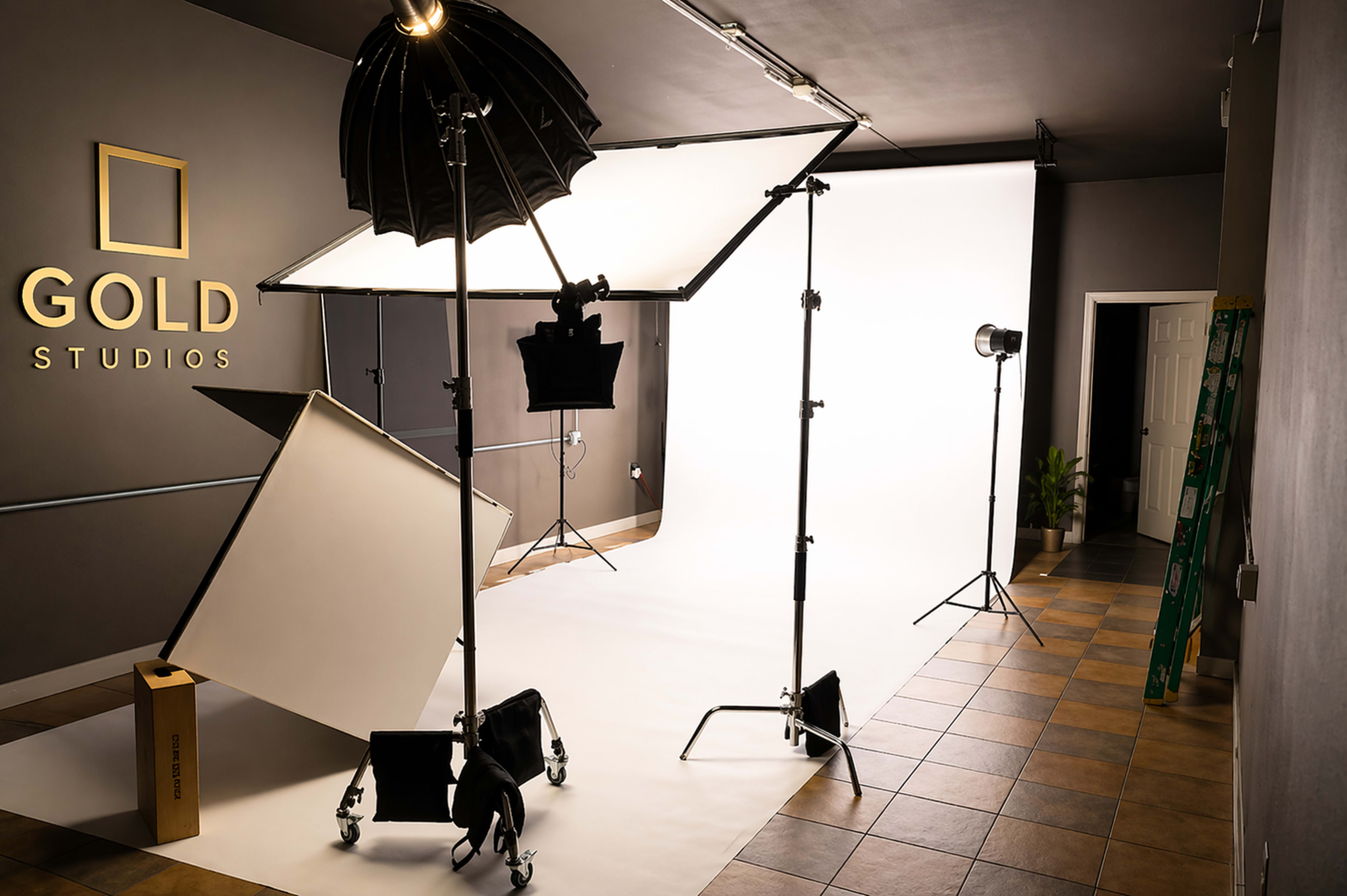 The image shows a photography studio setup with lighting equipment, backdrops, and a decorated wall featuring the name "GOLD STUDIOS."