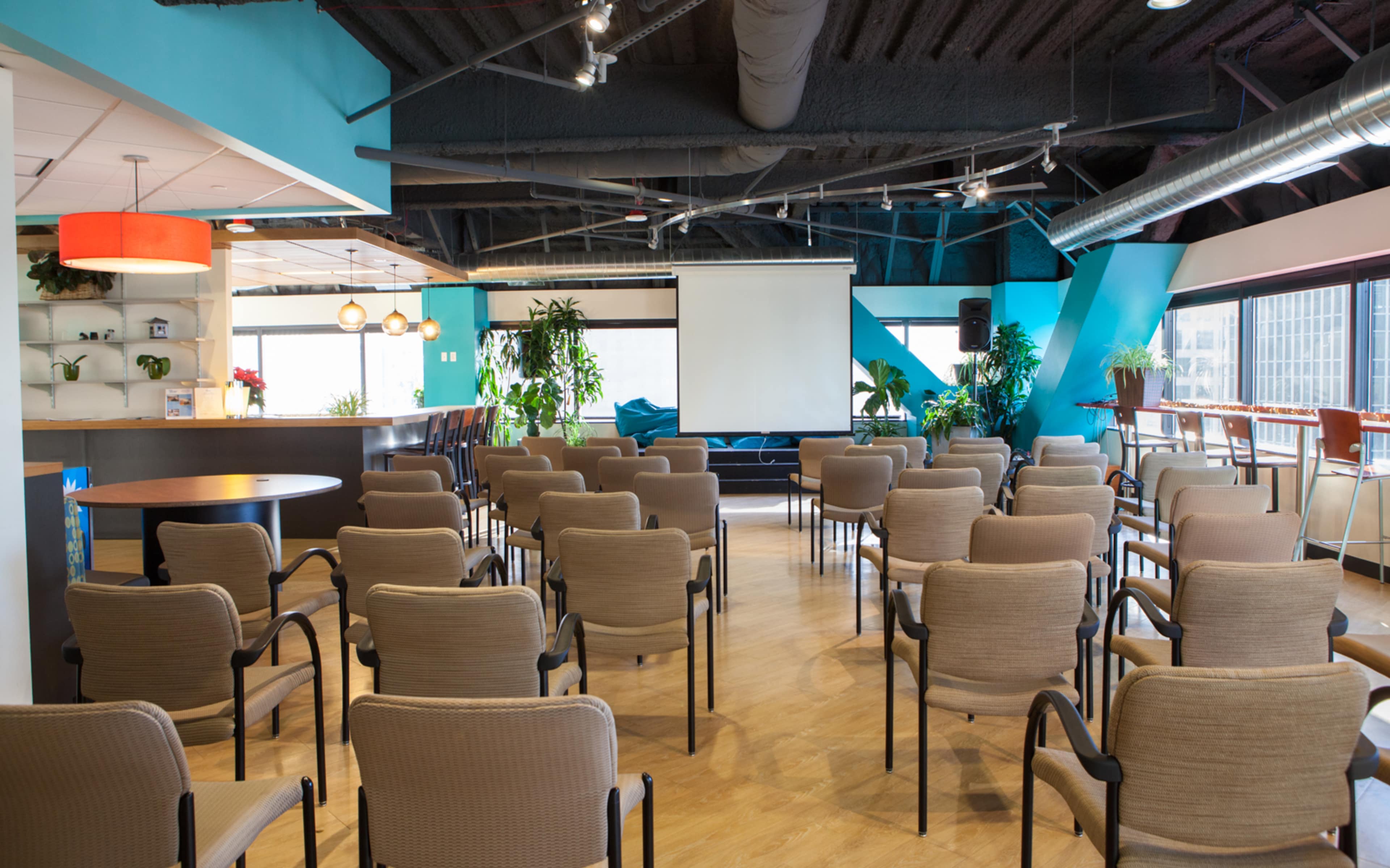 A conference room set up with rows of beige chairs facing a projection screen, accompanied by greenery and modern decor.