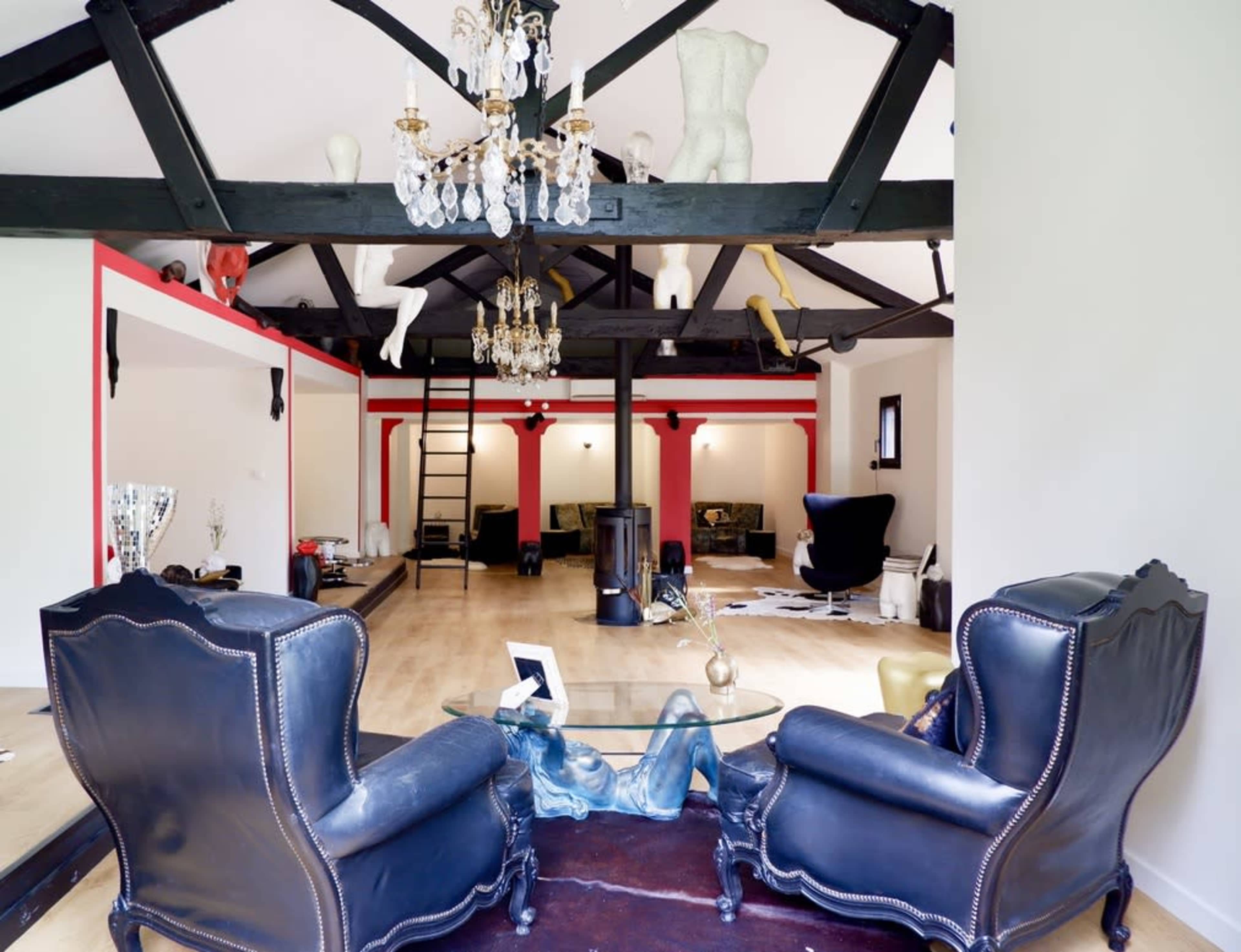 The image shows a spacious interior with black wooden beams, a chandelier, and a combination of black armchairs and modern sculptures, set against light-colored walls.