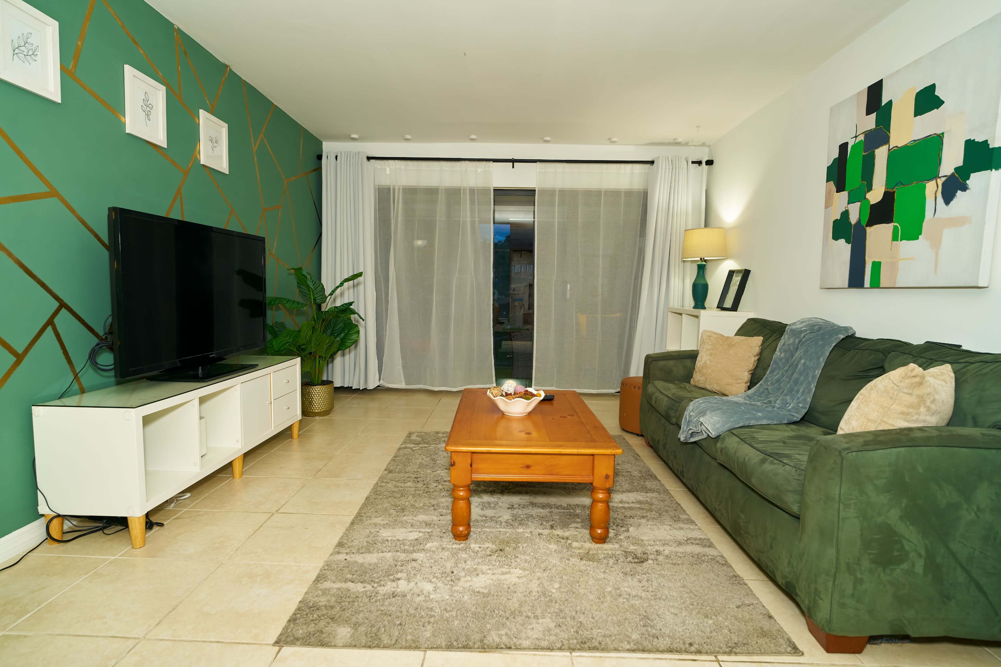 The image shows a living room with a green accent wall, a television on a white cabinet, a wooden coffee table, a green sofa, and large sliding glass doors with white curtains.