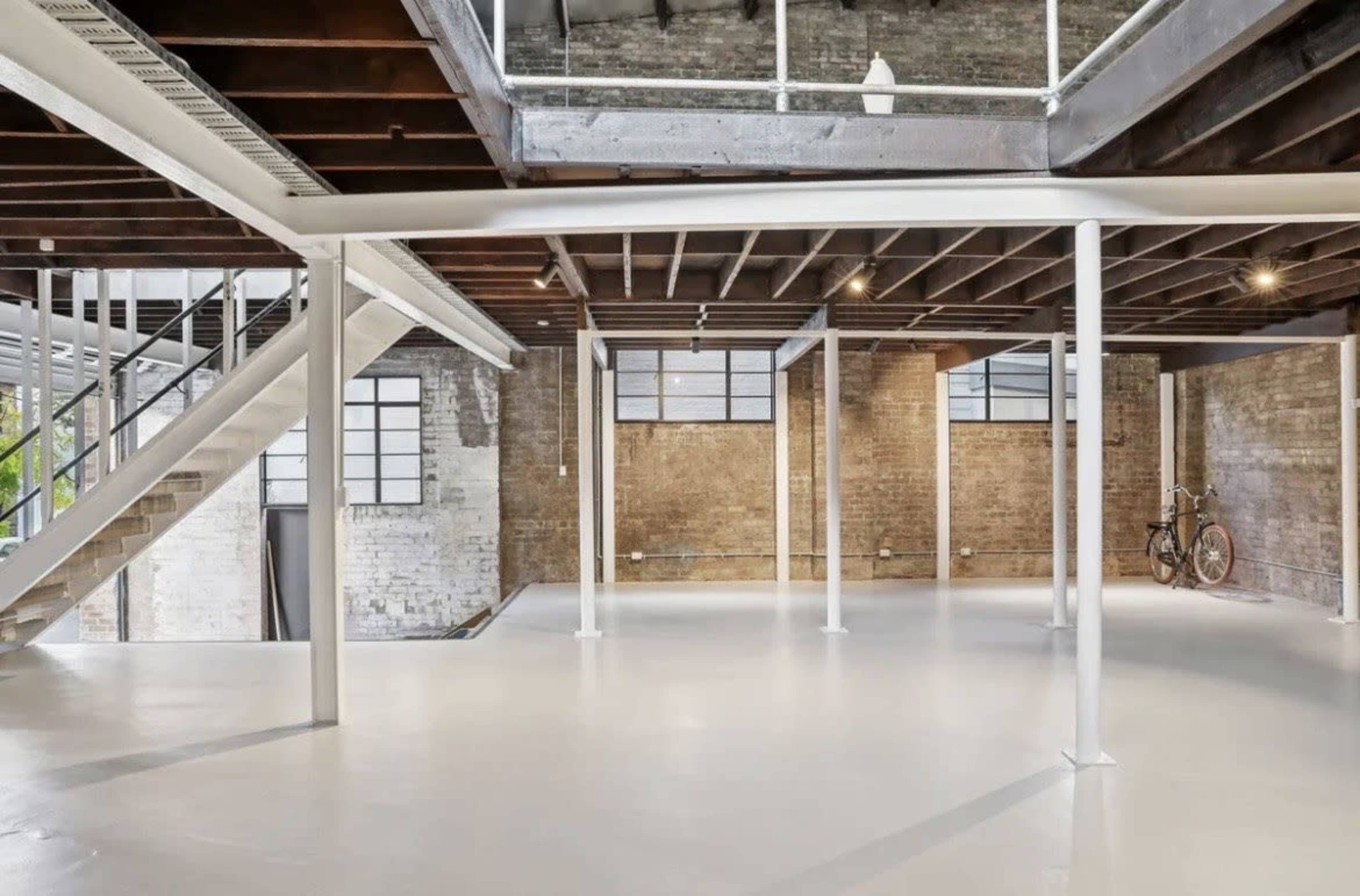 The image shows a spacious, industrial-style interior with exposed brick walls, a polished white floor, and large windows, featuring a staircase and a bicycle in the corner.
