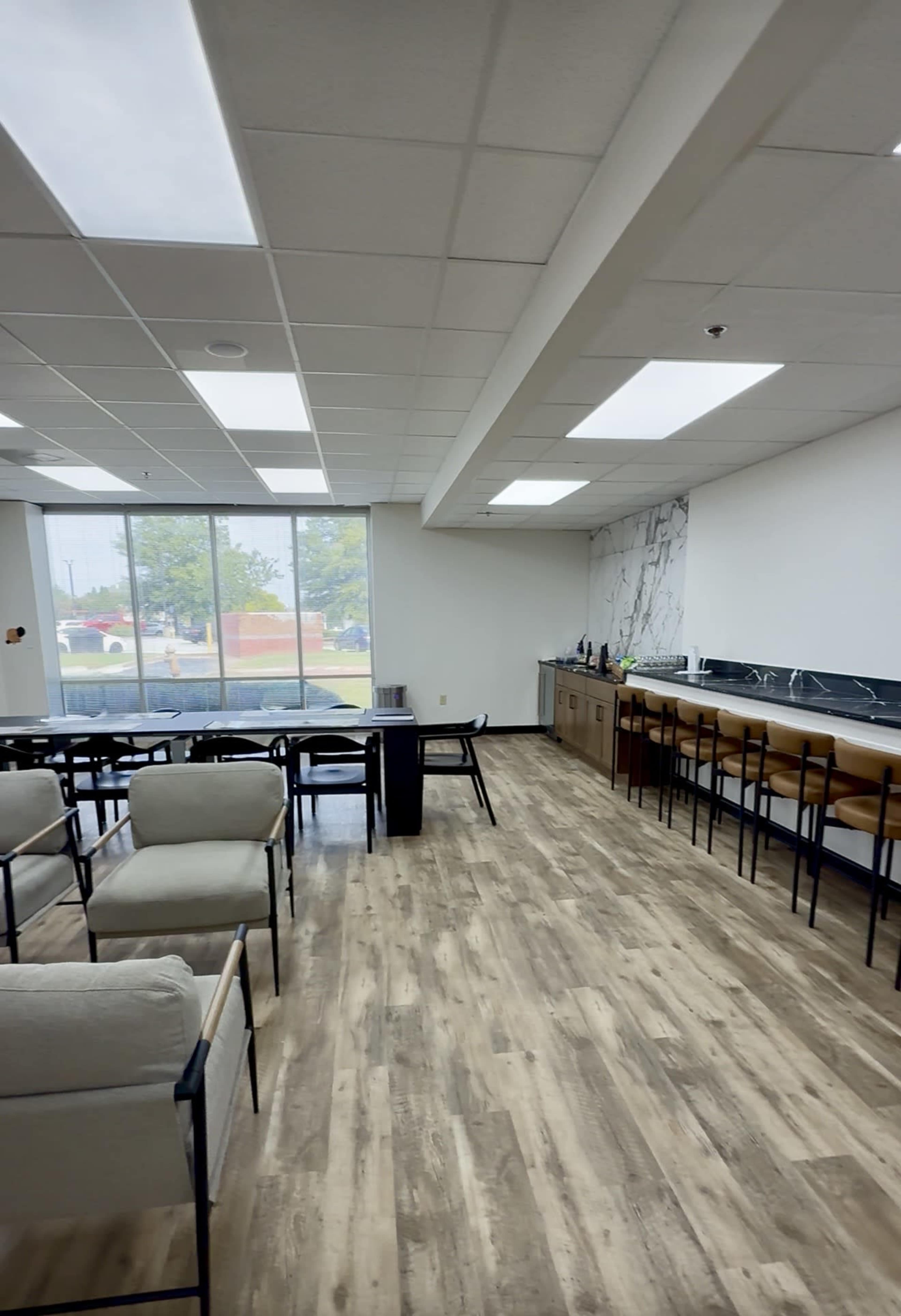 The image shows a modern, minimally furnished room with a seating area on one side and a bar counter on the other, featuring large windows that allow natural light to enter.