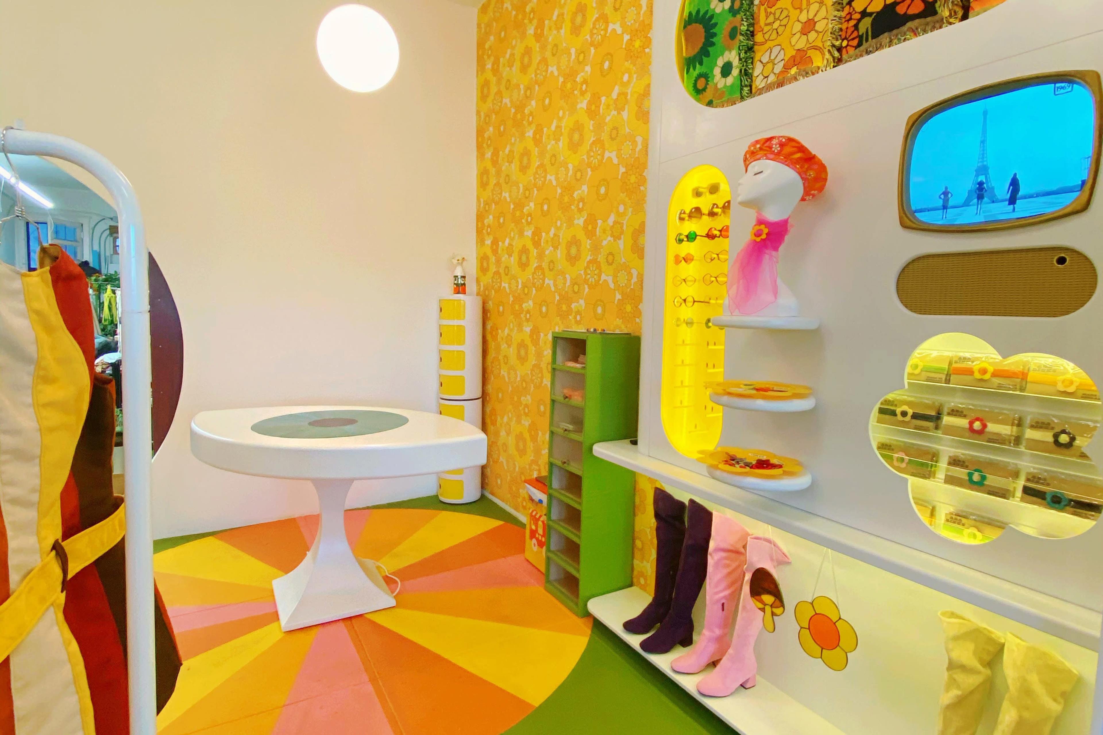 The image displays a colorful vintage-style boutique with a circular table, a wall of accessories, and garments displayed on white stands.