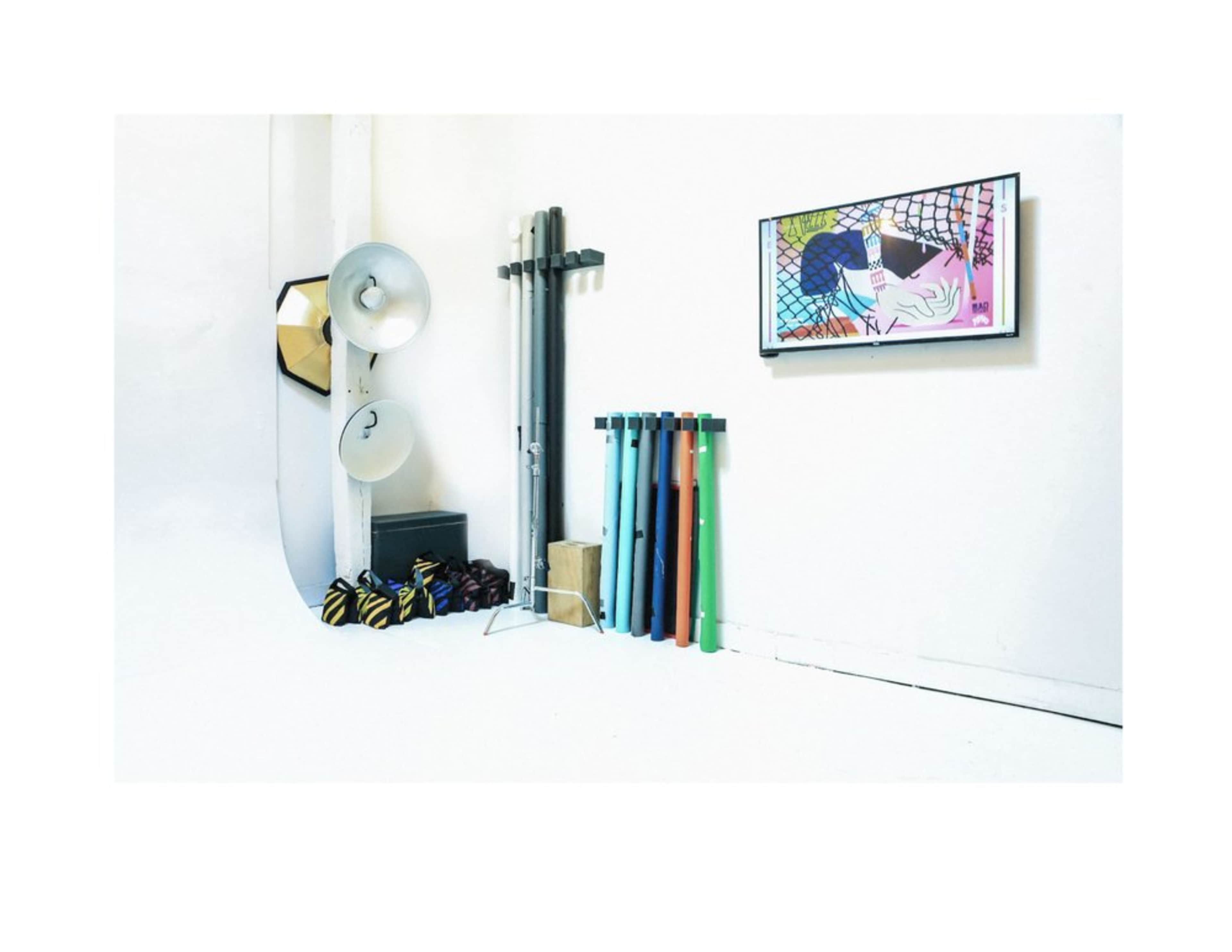 The image shows a minimalist studio space featuring several colorful poles, lighting equipment, and a wall-mounted artwork.