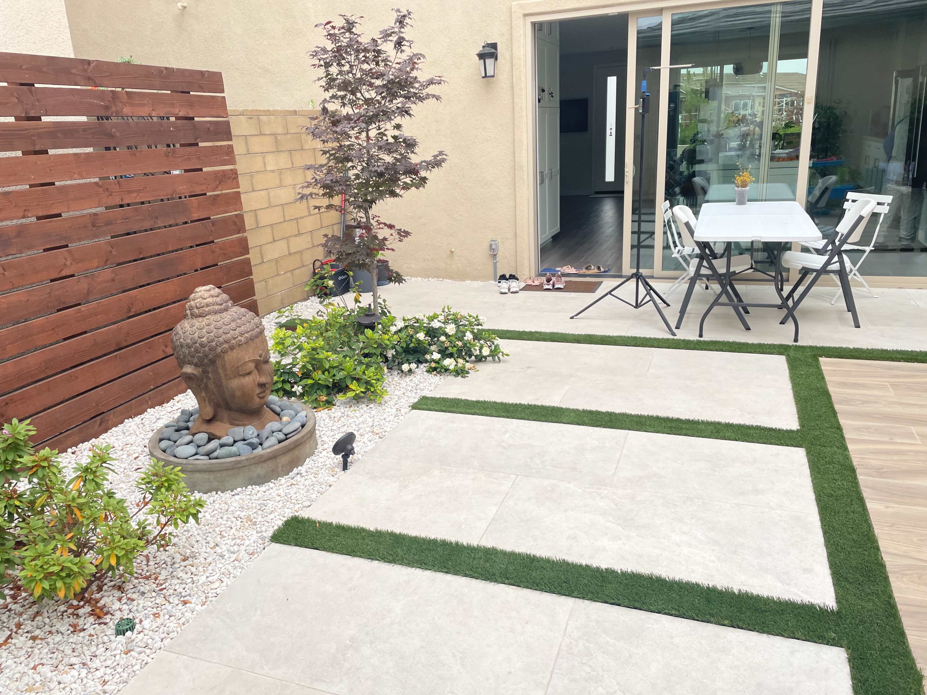 A patio area featuring a stone Buddha statue, bordered by plants and decorative gravel, with a small table and chairs visible through a nearby sliding glass door.