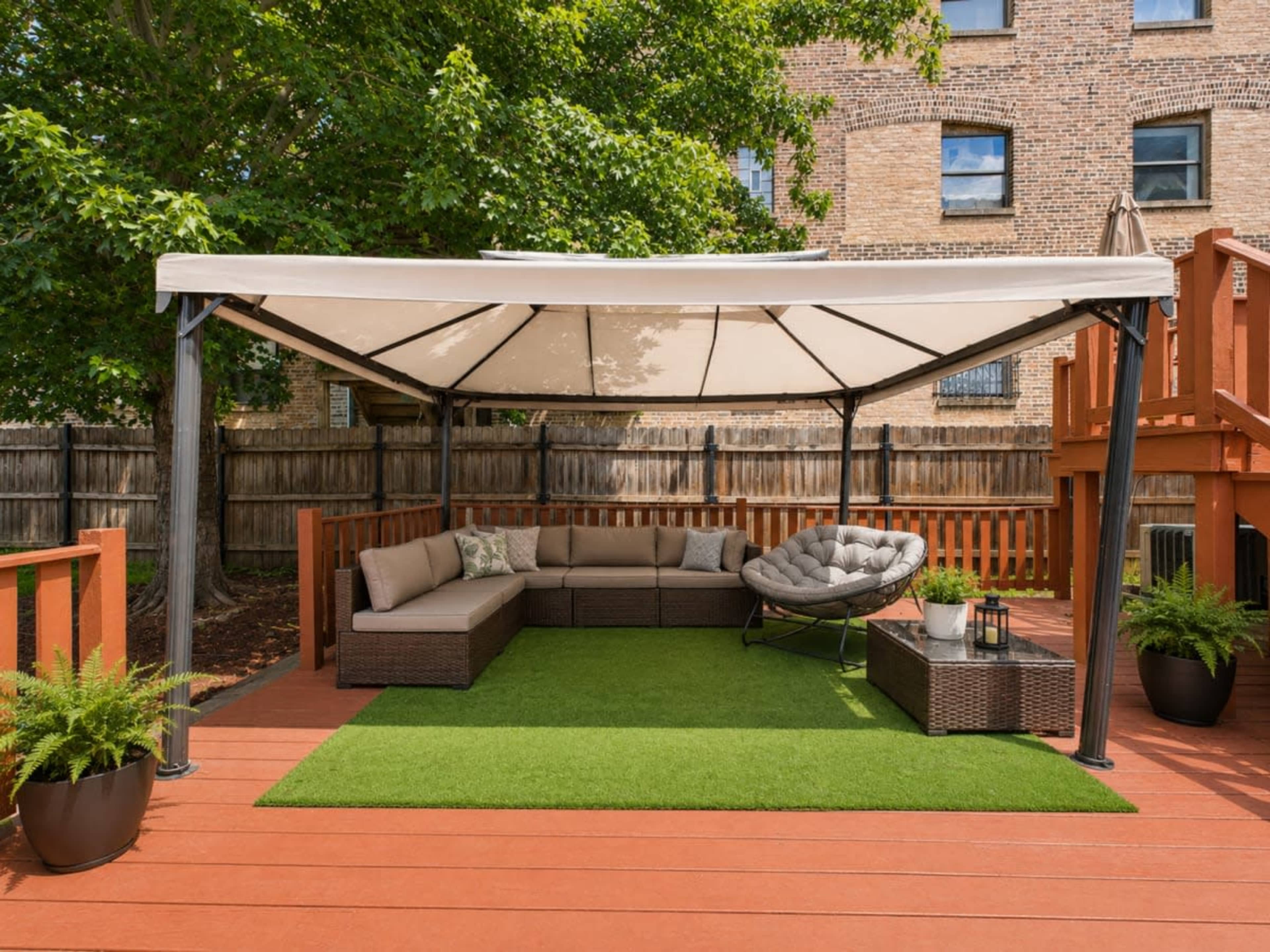 The image shows a shaded outdoor seating area with a sectional sofa, a circular chair, and a small table on a green turf surface, surrounded by wooden fencing and trees.