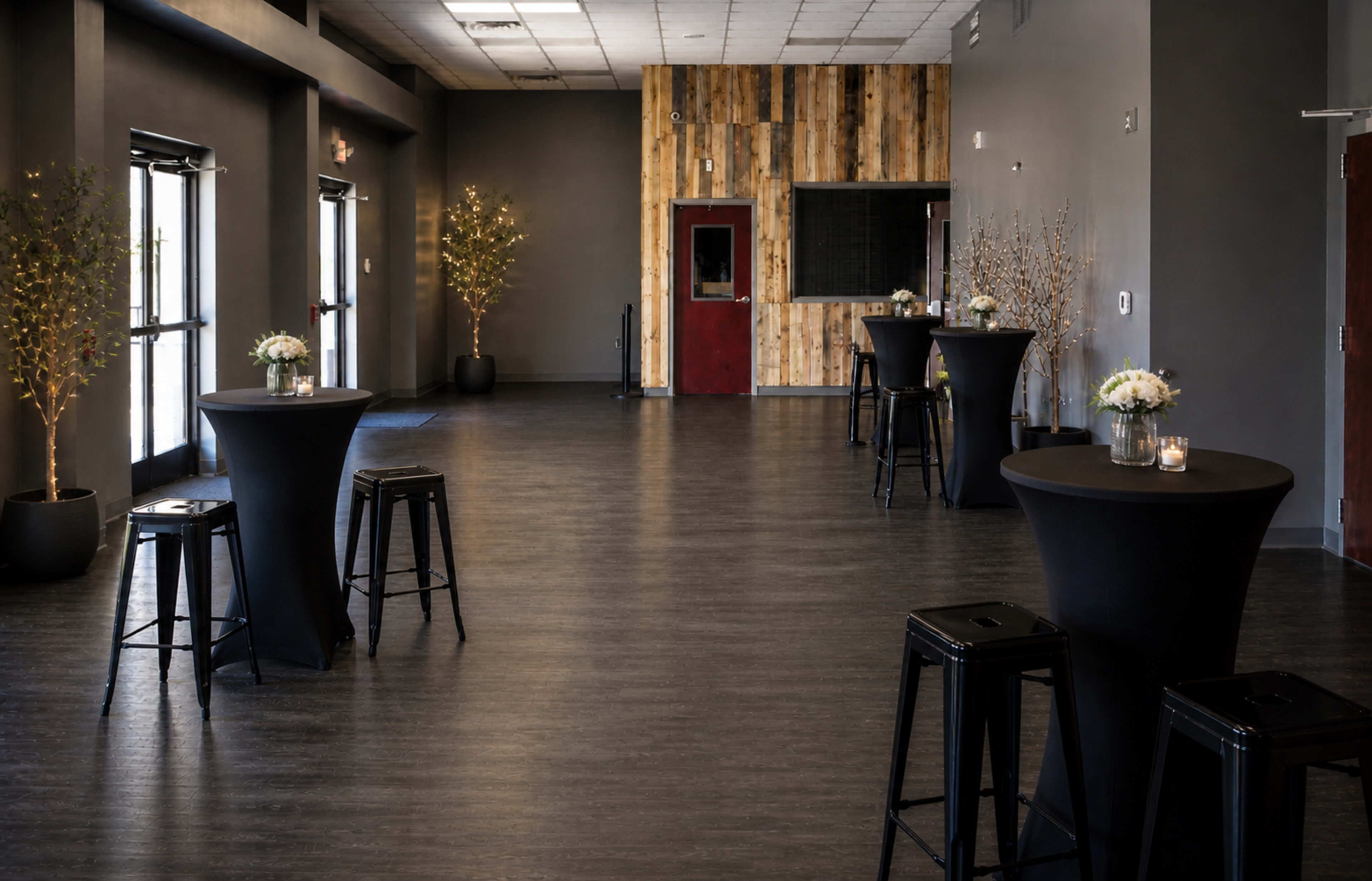 The image shows an event space with several tall black tables surrounded by bar stools, illuminated by soft lighting and featuring wooden accents on the wall.