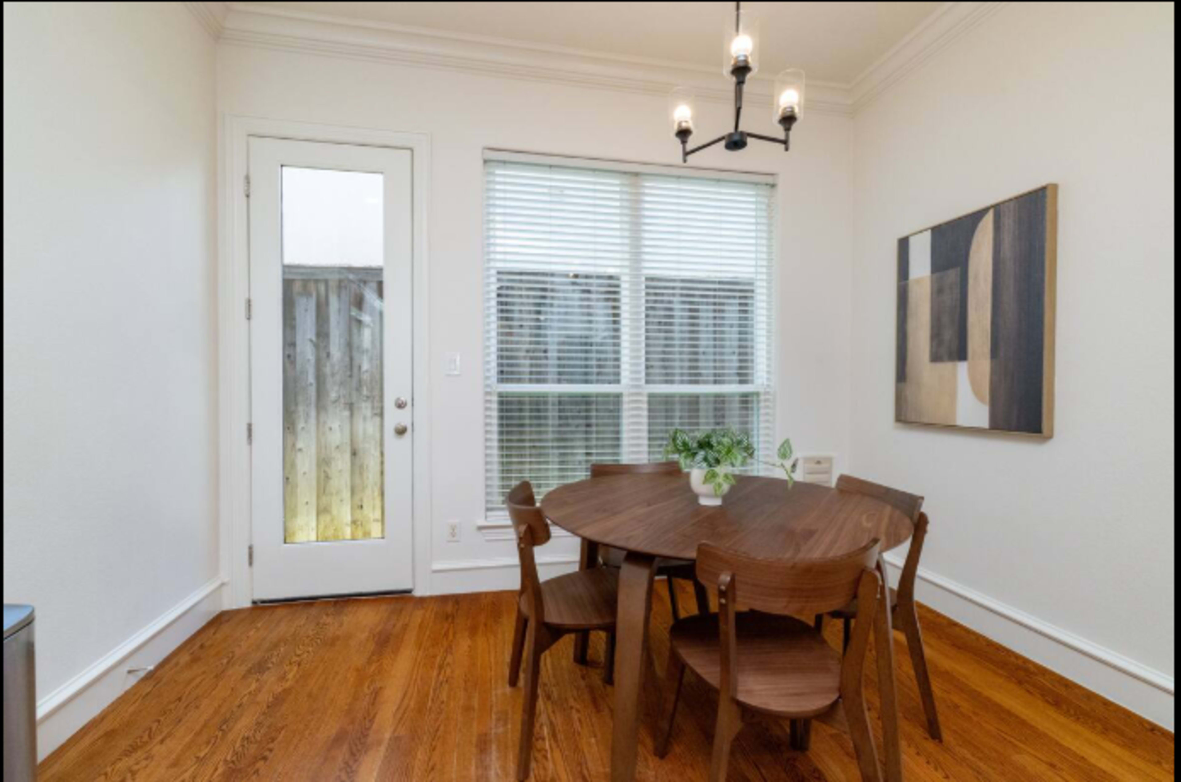 A round wooden dining table surrounded by four chairs is positioned near a large window with blinds, with a door leading outside in the background.