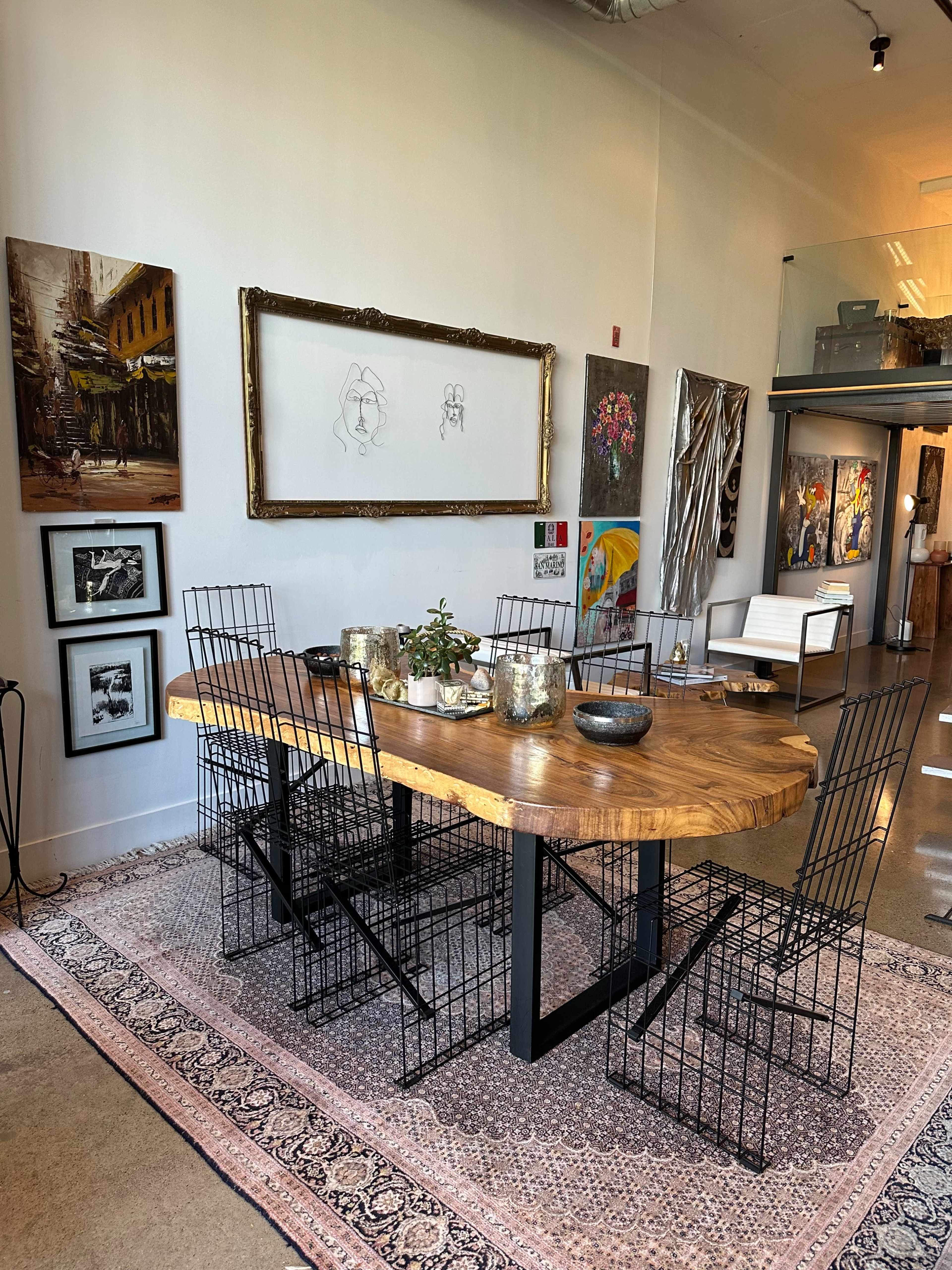A large wooden dining table is set with dishes and surrounded by black wire chairs, located in an art-filled room with various artworks on the walls.