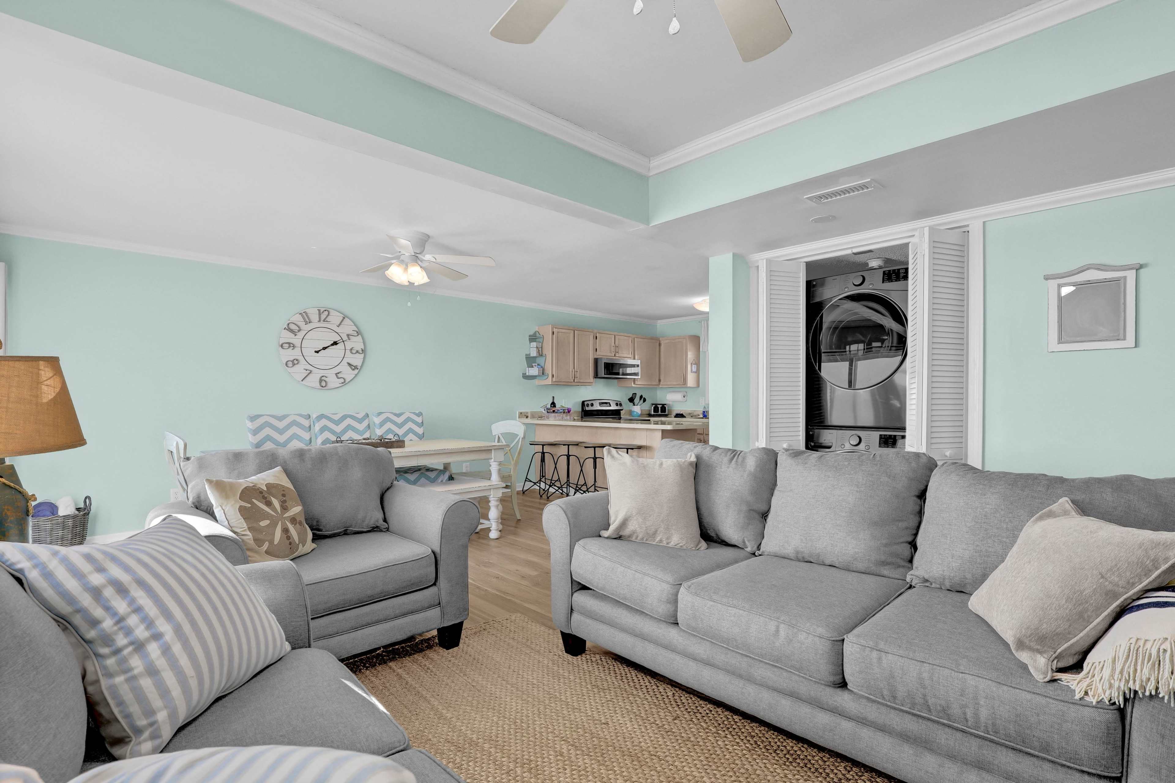 The image shows a light-colored living room with two gray sofas, a wall clock, and a kitchen area visible in the background.