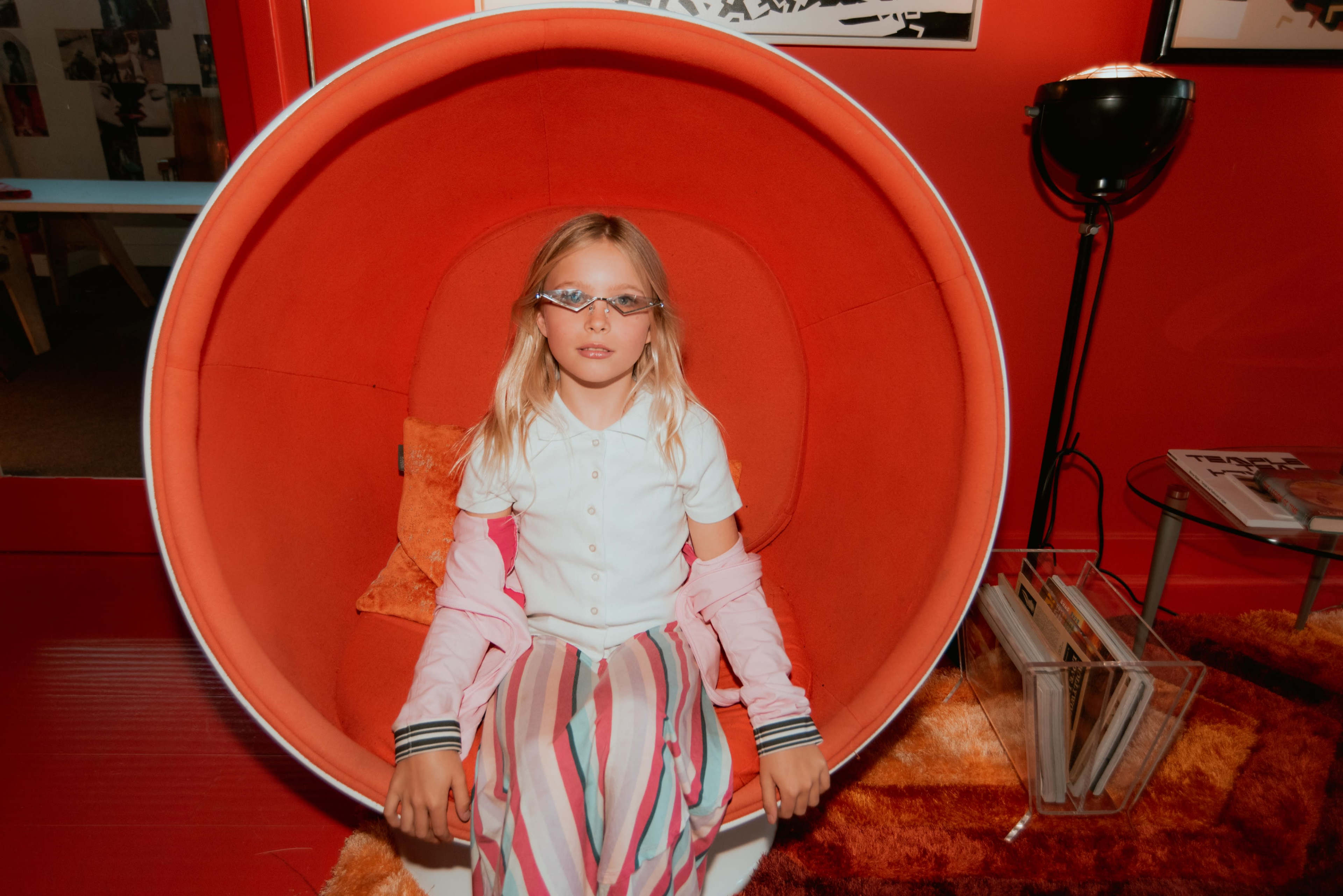A girl with glasses sits in a round orange chair in a room with red walls and various decor.