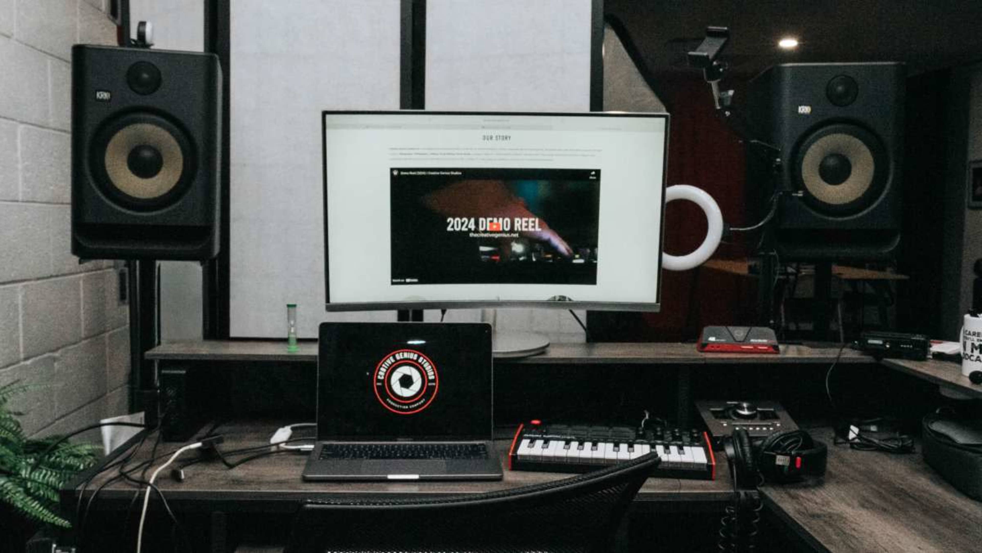 A music production workspace features a computer monitor displaying a demo reel, a laptop with a keyboard, and large speakers on either side.
