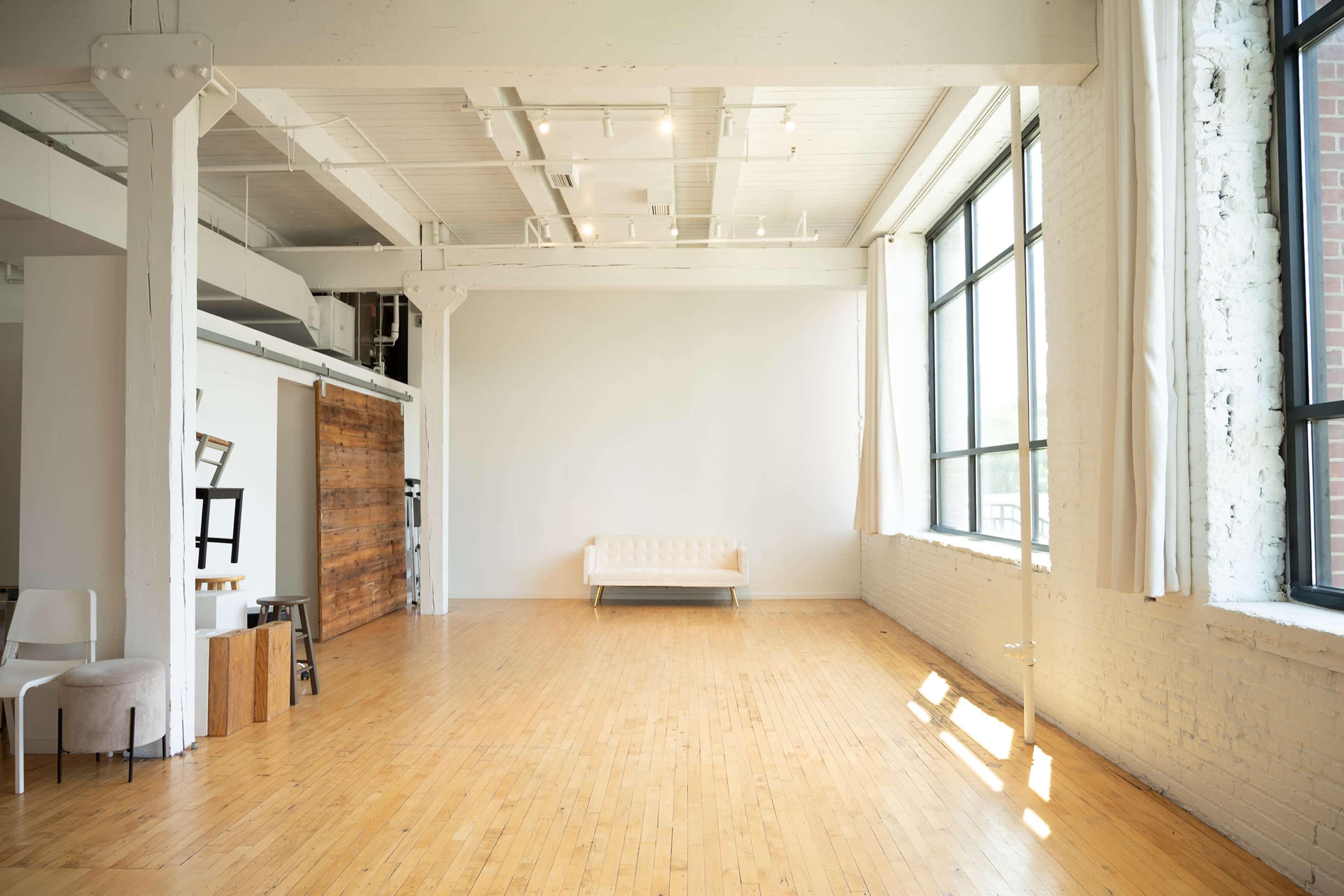The image shows a spacious, bright open room with wooden flooring, large windows, and minimal furnishings, including a sofa and a few chairs.