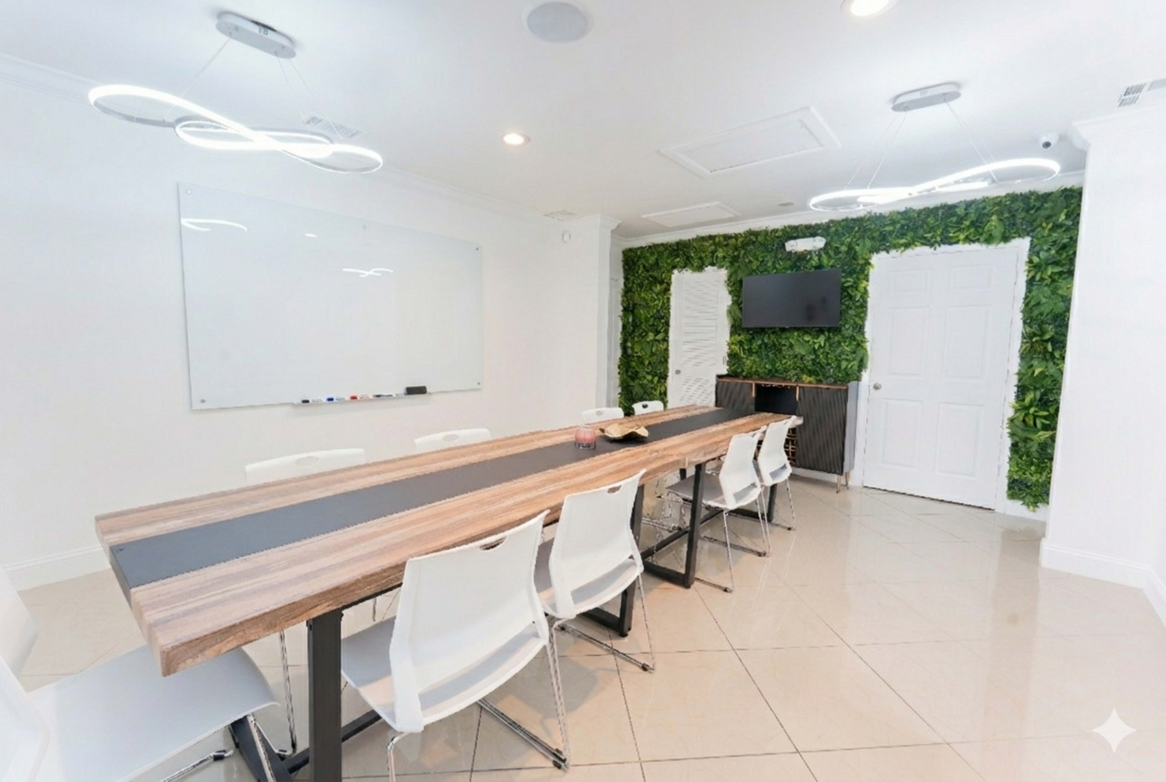 A modern conference room features a long wooden table, white chairs, a whiteboard, a television, and a green wall with plants.