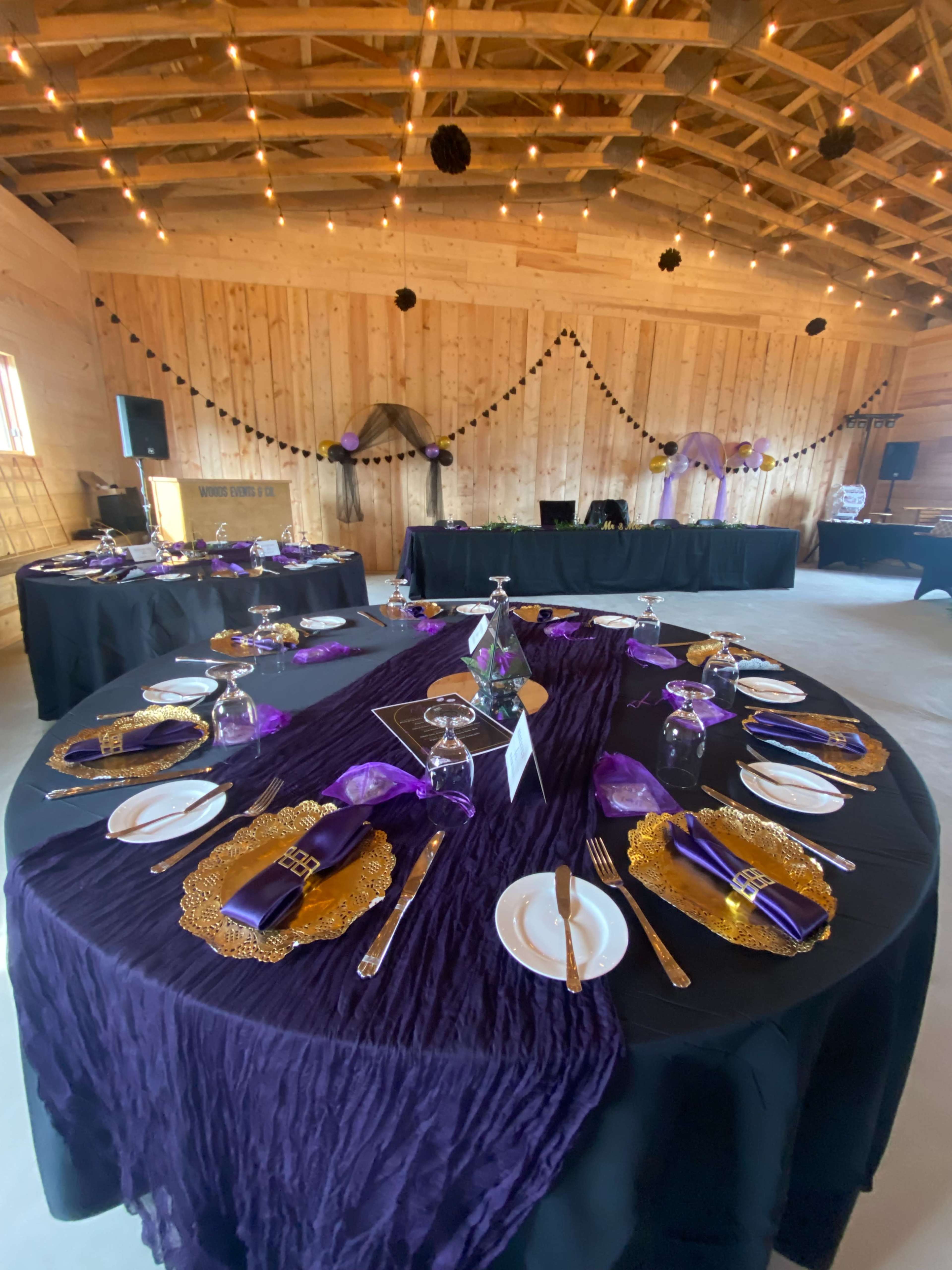 A banquet hall is elegantly set with round tables draped in purple and black tablecloths, adorned with golden chargers, crystal glassware, and decorative centerpieces, all under string lights.