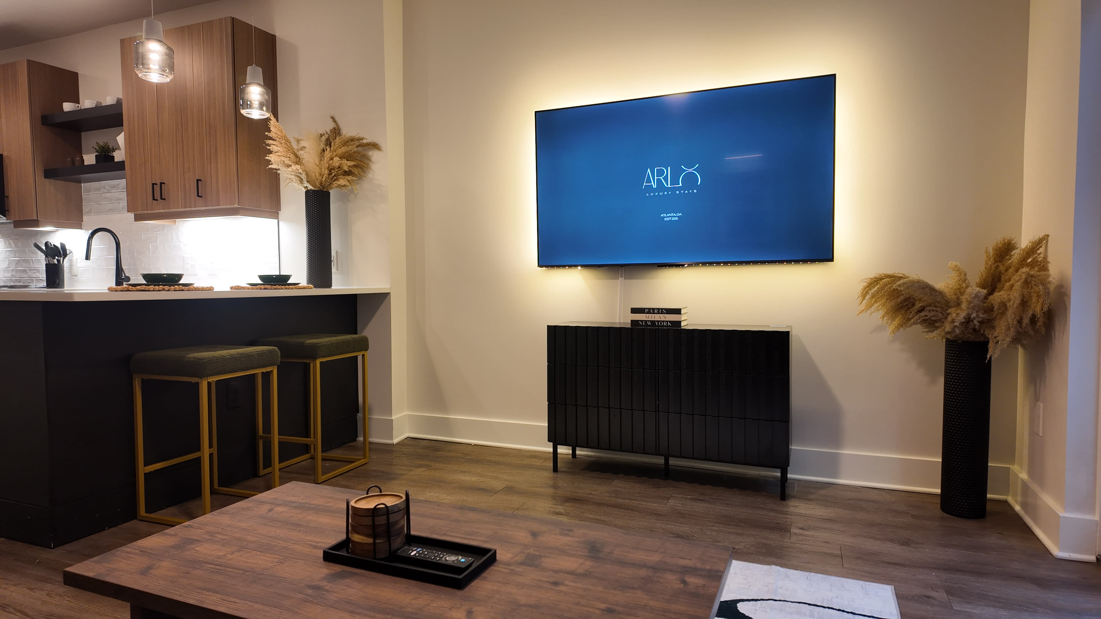 A modern living space features a wall-mounted TV above a sleek black cabinet, with a kitchen area in the background and decorative plants in tall vases flanking the TV.