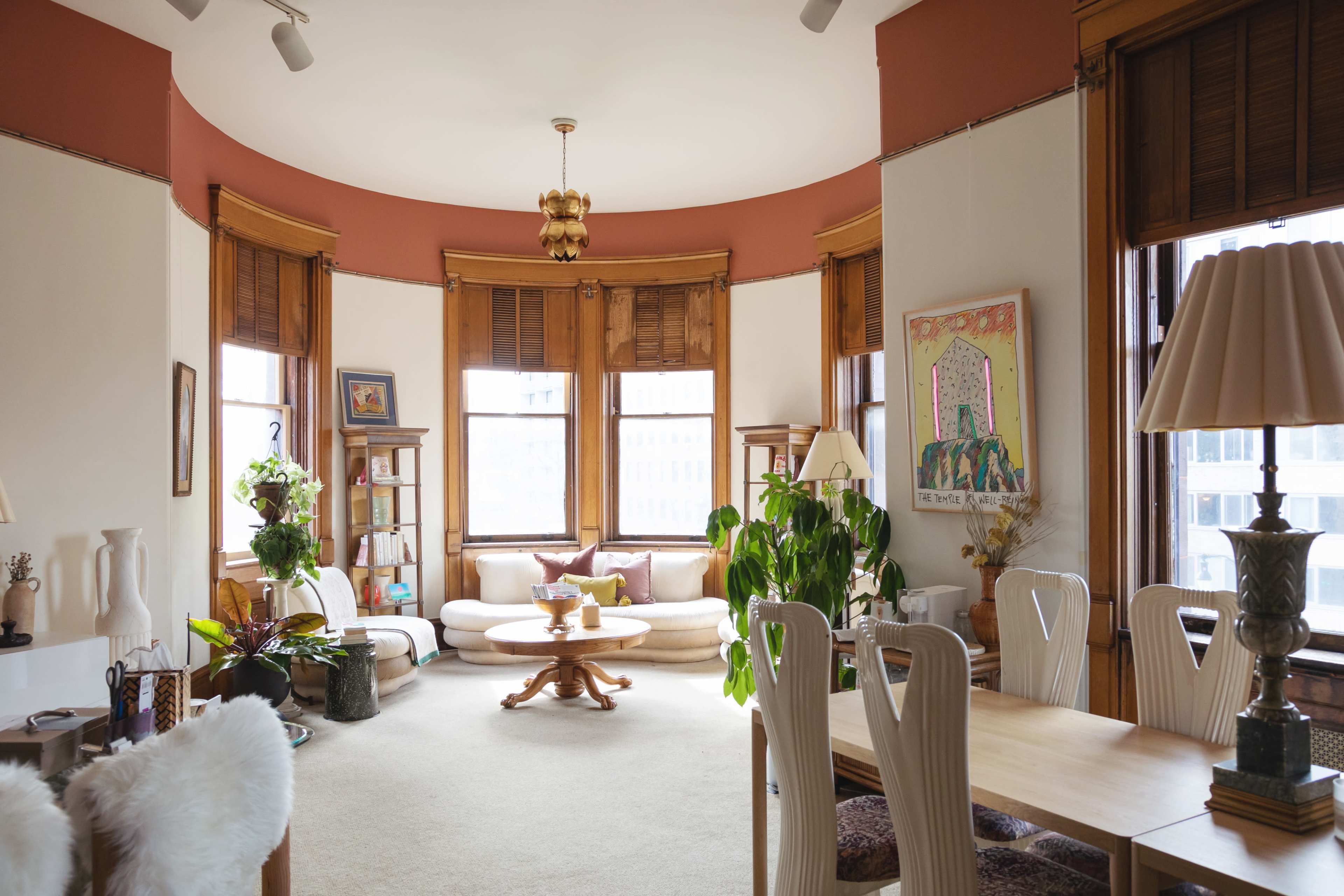A spacious living area with light-colored walls, large windows, and a circular arrangement of furniture, including a dining table and various plants.
