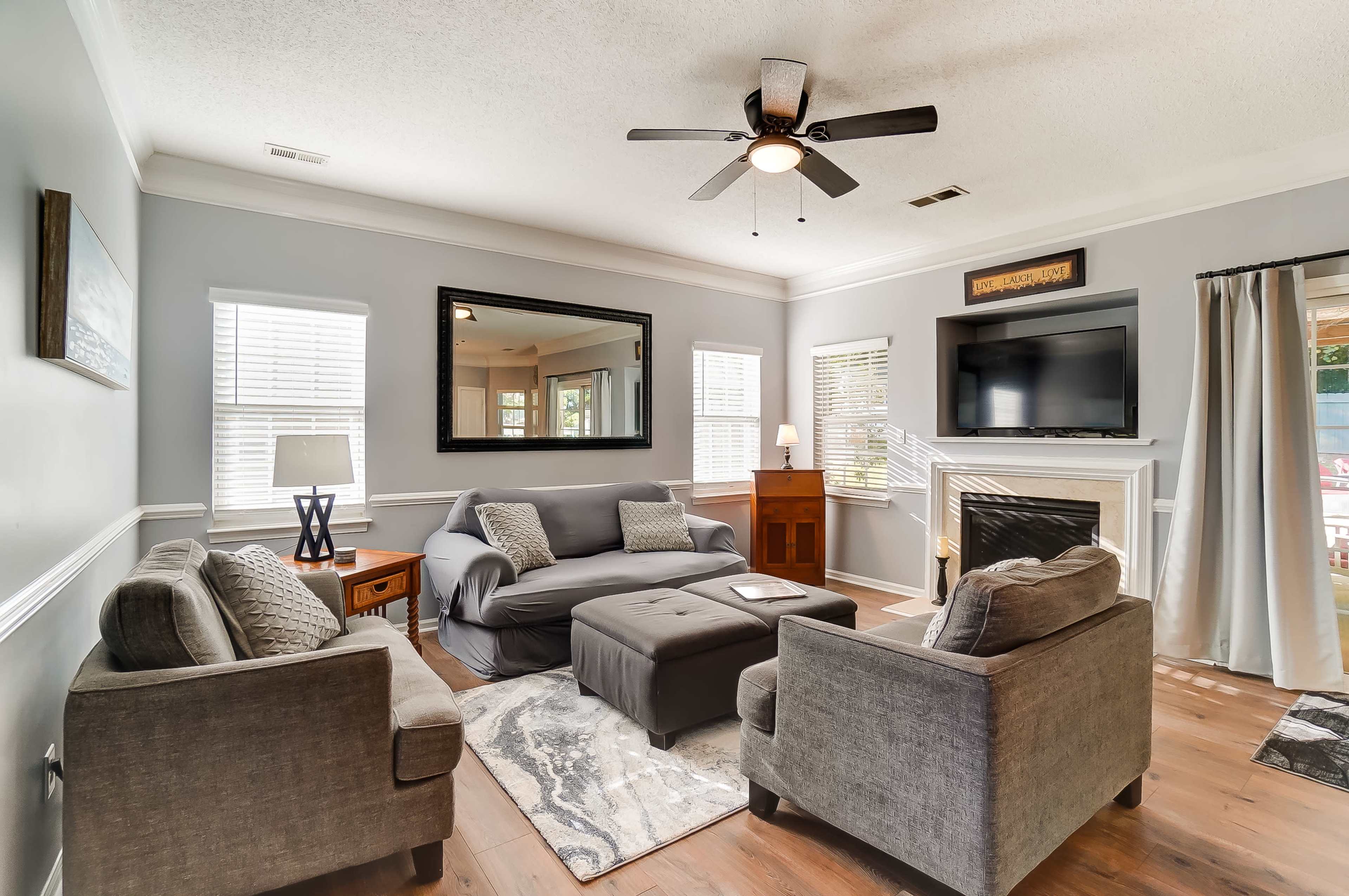 A living room features a gray sofa set, a coffee table, a wall-mounted television, and a fireplace, with large windows allowing natural light.