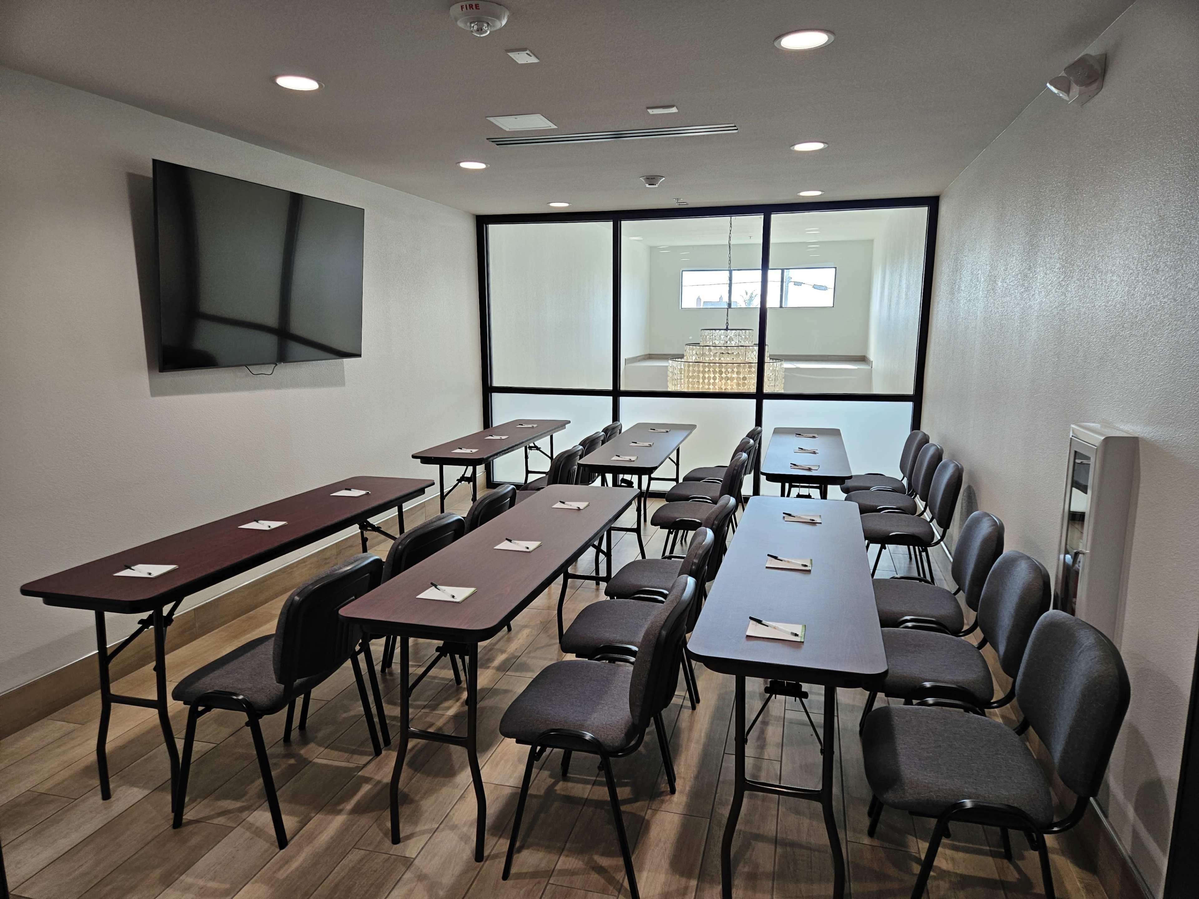 A small conference room is set up with several tables arranged in a U-shape and chairs positioned around them, with a large screen mounted on one wall.