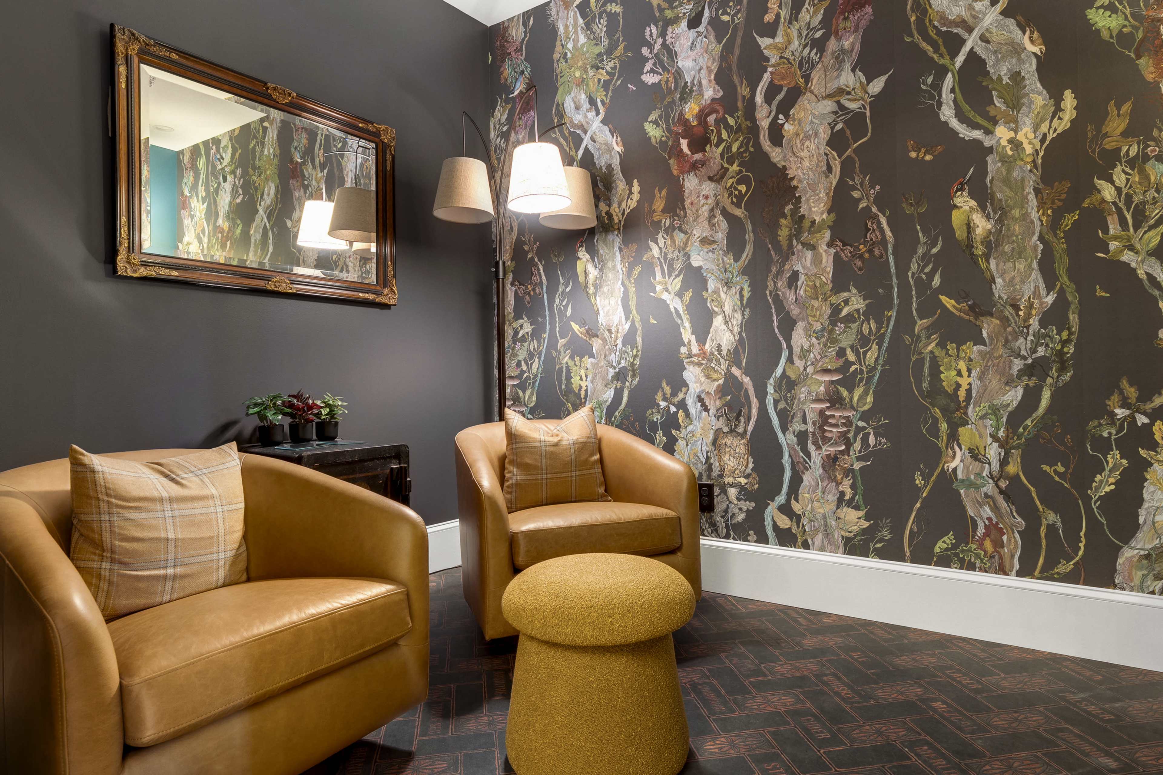 The image features a cozy seating area with two brown leather chairs, a small round yellow table, and a decorative wall covered in a floral-patterned wallpaper.