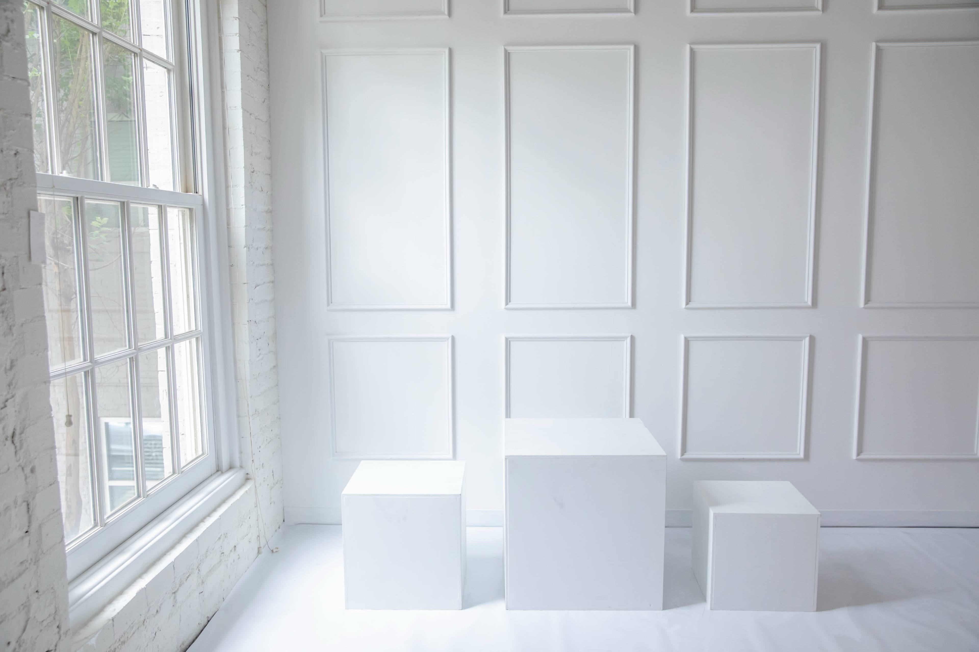 The image shows three white cubes of varying heights arranged against a white-paneled wall, with a window providing natural light.