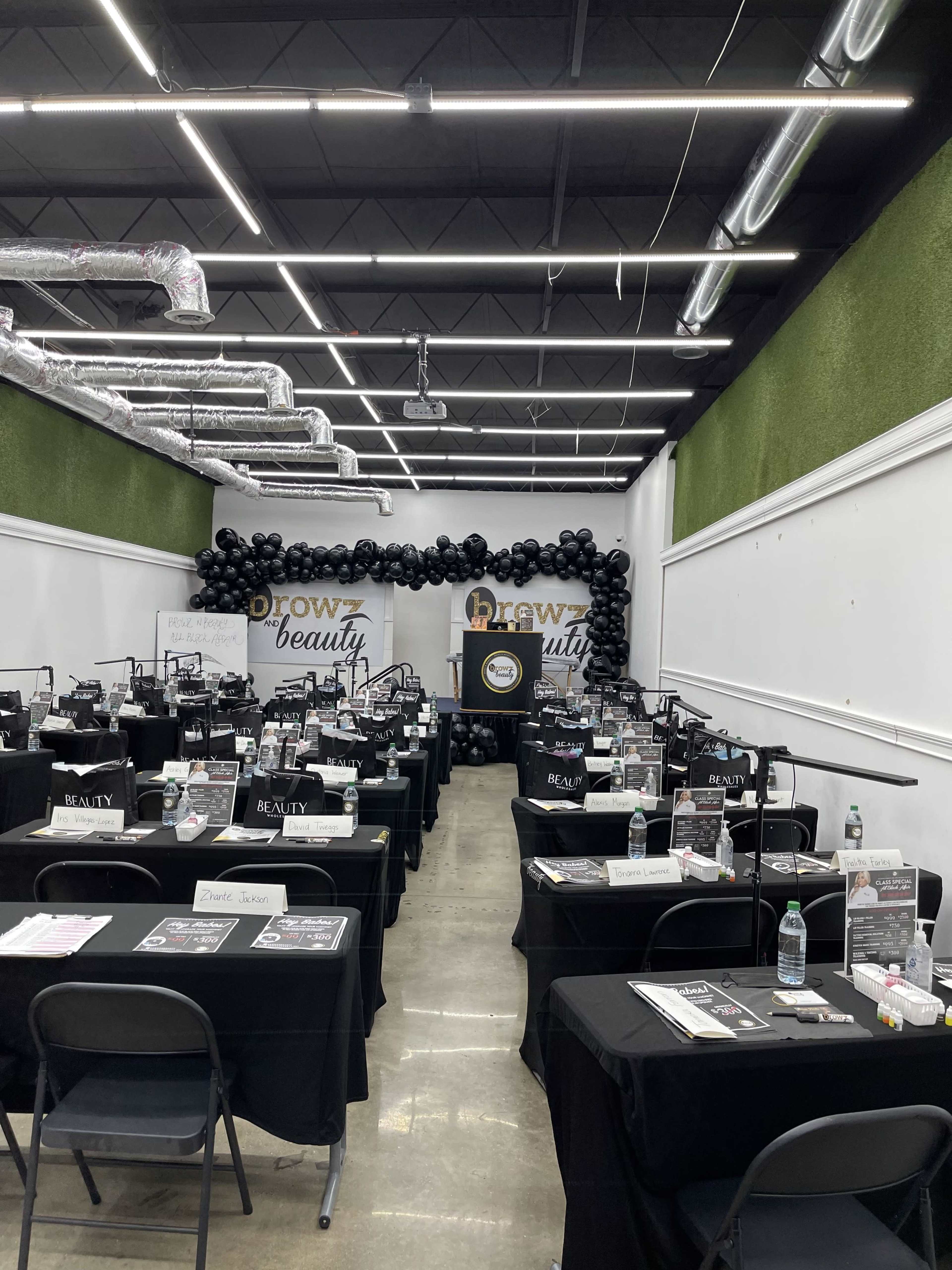 A classroom or event space set up with multiple tables covered in black cloth, each equipped with beauty supplies and tools, arranged in rows under black balloon decorations.