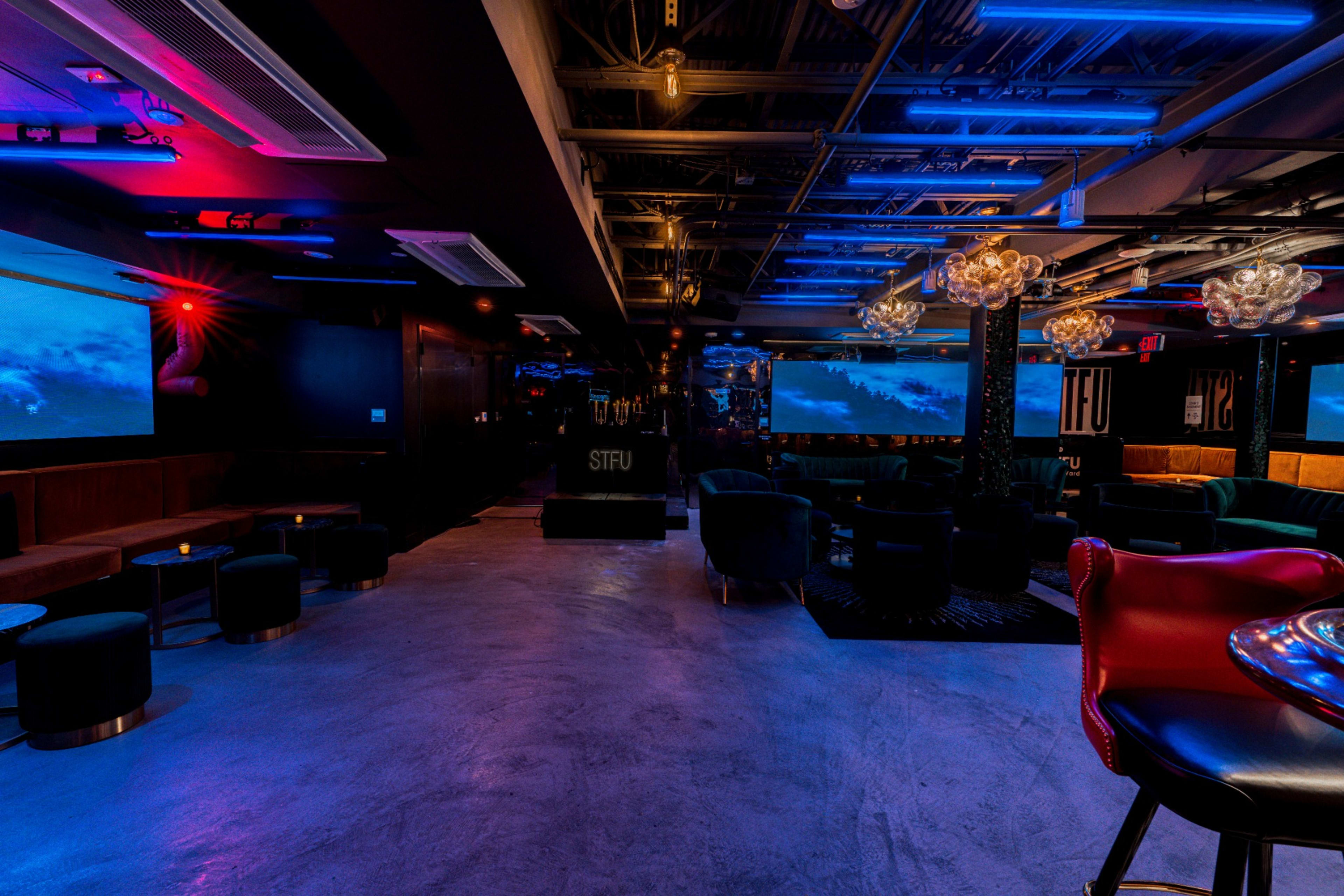 The interior of a modern lounge features dark seating, colorful neon lights, and large screens displaying visual content.