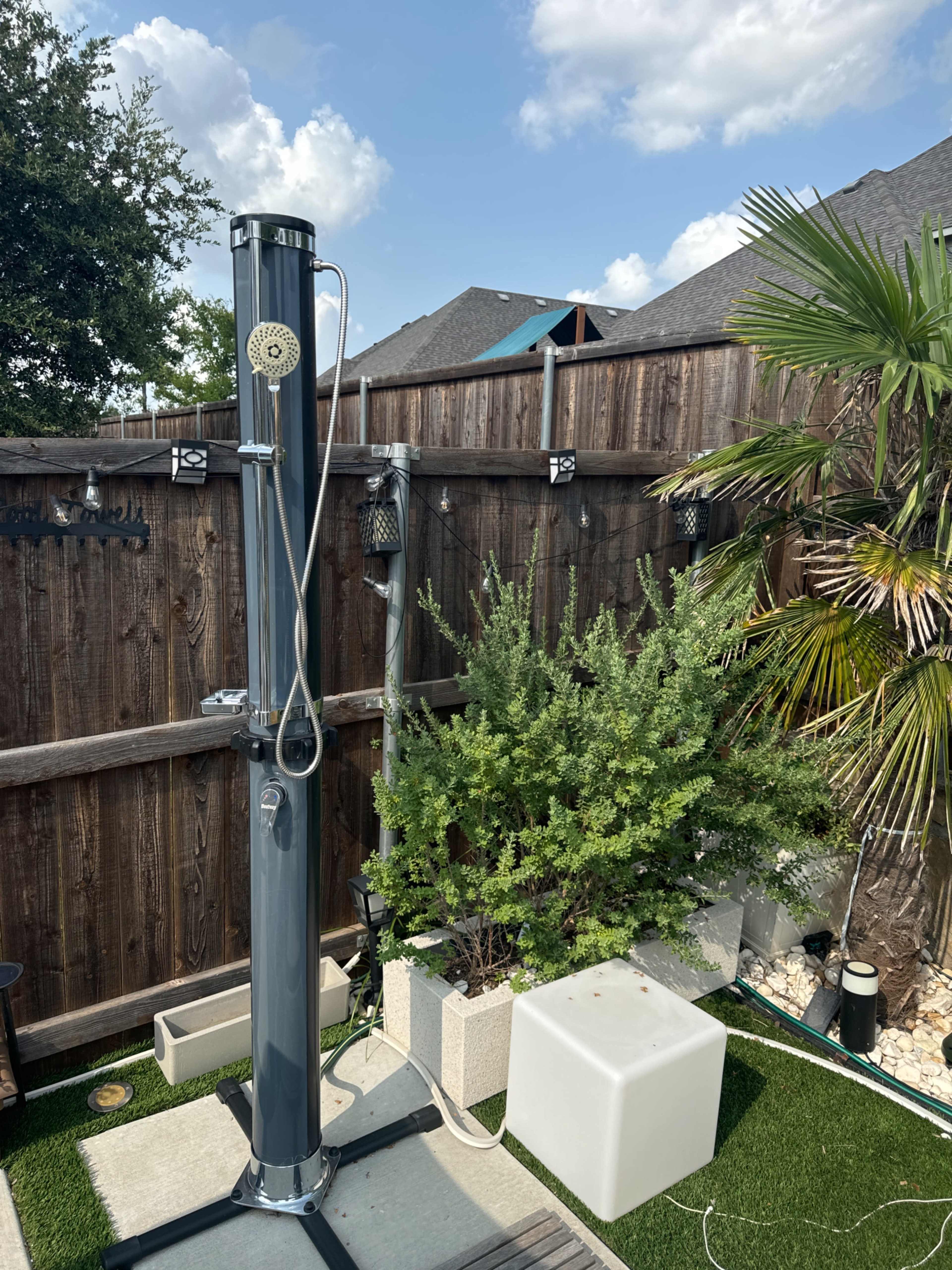 An outdoor shower mounted on a wooden fence next to a planted area with shrubs and a small white stool.